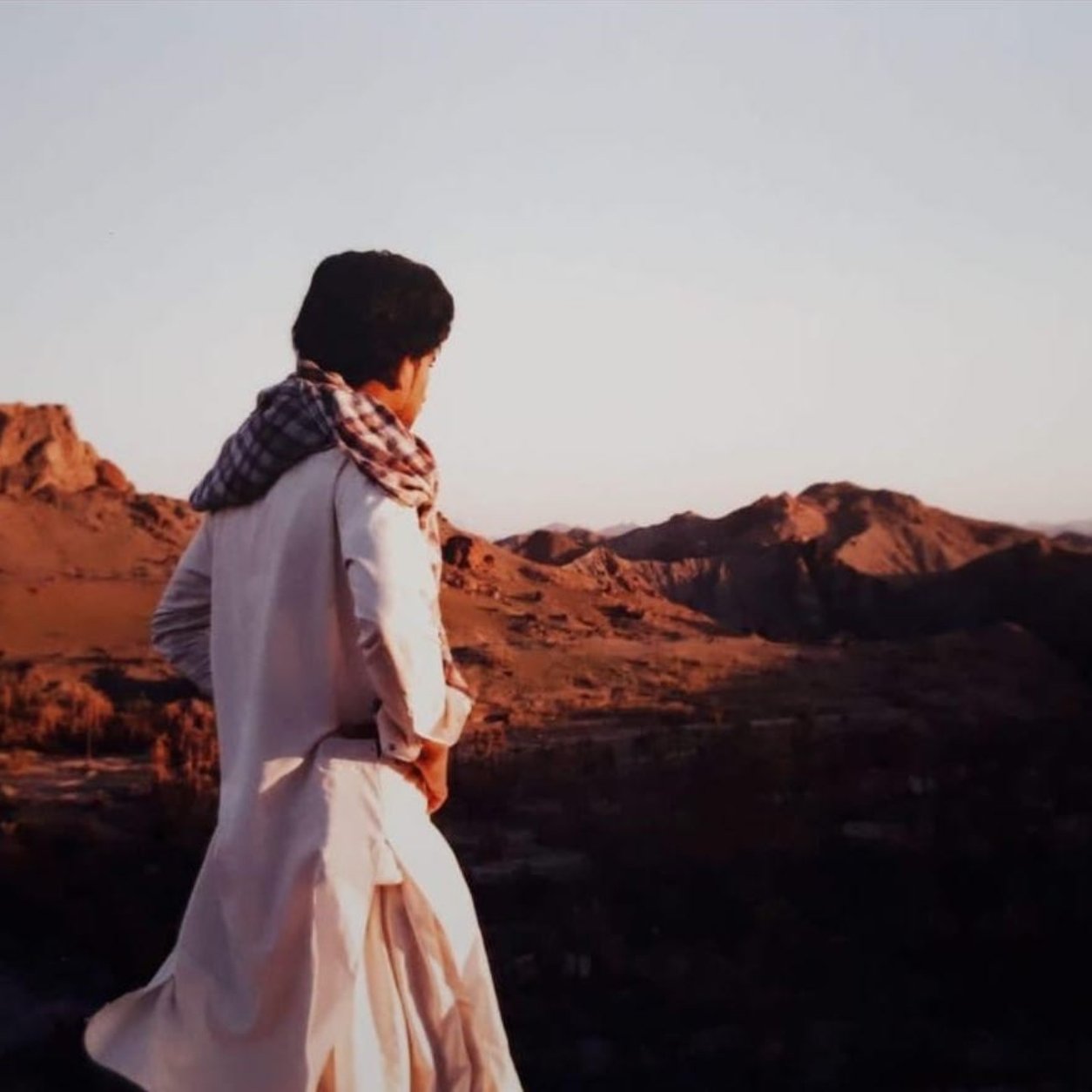L'image montre une personne debout sur une colline, regardant au loin vers un paysage montagneux. La lumière douce du coucher de soleil illumine les montagnes environnantes, créant une atmosphère sereine. La personne porte une tenue traditionnelle, avec un chèche sur la tête, qui évoque une culture spécifique. L'arrière-plan est composé de collines et de paysages arides, typiques de certaines régions désertiques.