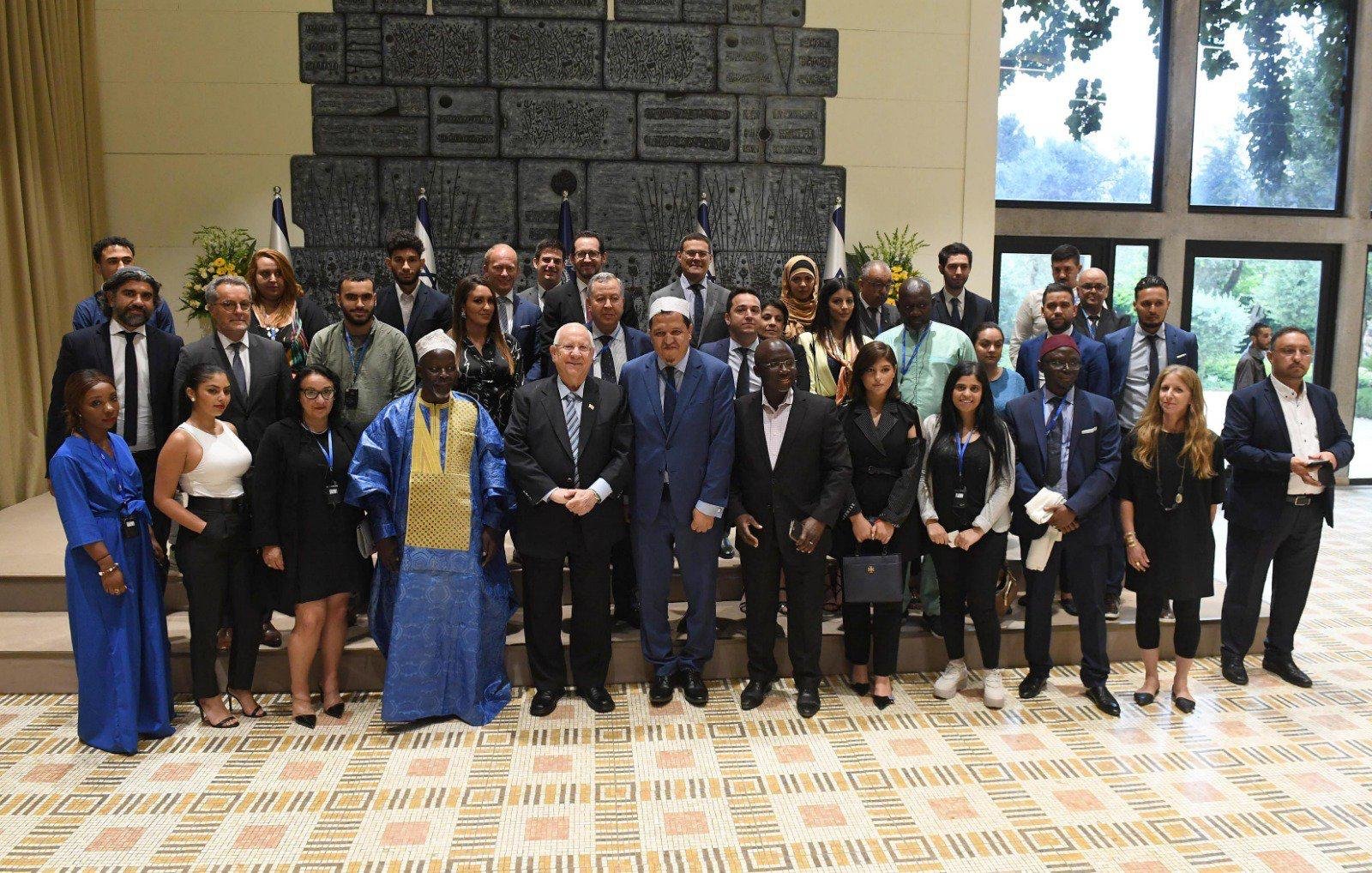 The image shows a large group of people gathered together for a formal event. They are standing in a room with a decorative backdrop that includes flags and a large, textured wall. The group consists of men and women of diverse backgrounds, and many are dressed in professional attire. Some individuals wear traditional cultural clothing. Everyone appears to be smiling, and they are posed together for a photograph, likely to commemorate the occasion. The setting is bright and well-lit, contributing to a positive atmosphere.