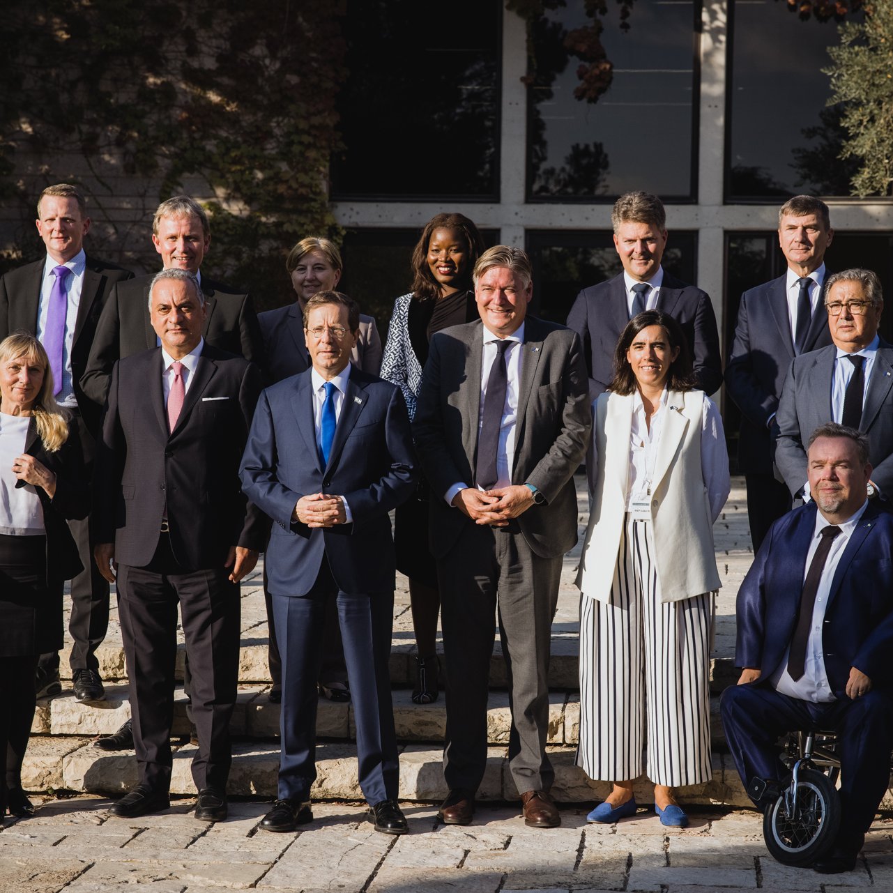 L'image montre un groupe de personnes posant ensemble à l'extérieur, sur des marches devant un bâtiment. Il y a dix-sept individus, dont certains portent des costumes et d'autres des tenues variées. L'arrière-plan est un jardin ou un espace extérieur avec des arbres et une architecture moderne. Les personnes affichent des expressions souriantes, ce qui suggère un moment de camaraderie ou une occasion formelle, comme une réunion ou un sommet.