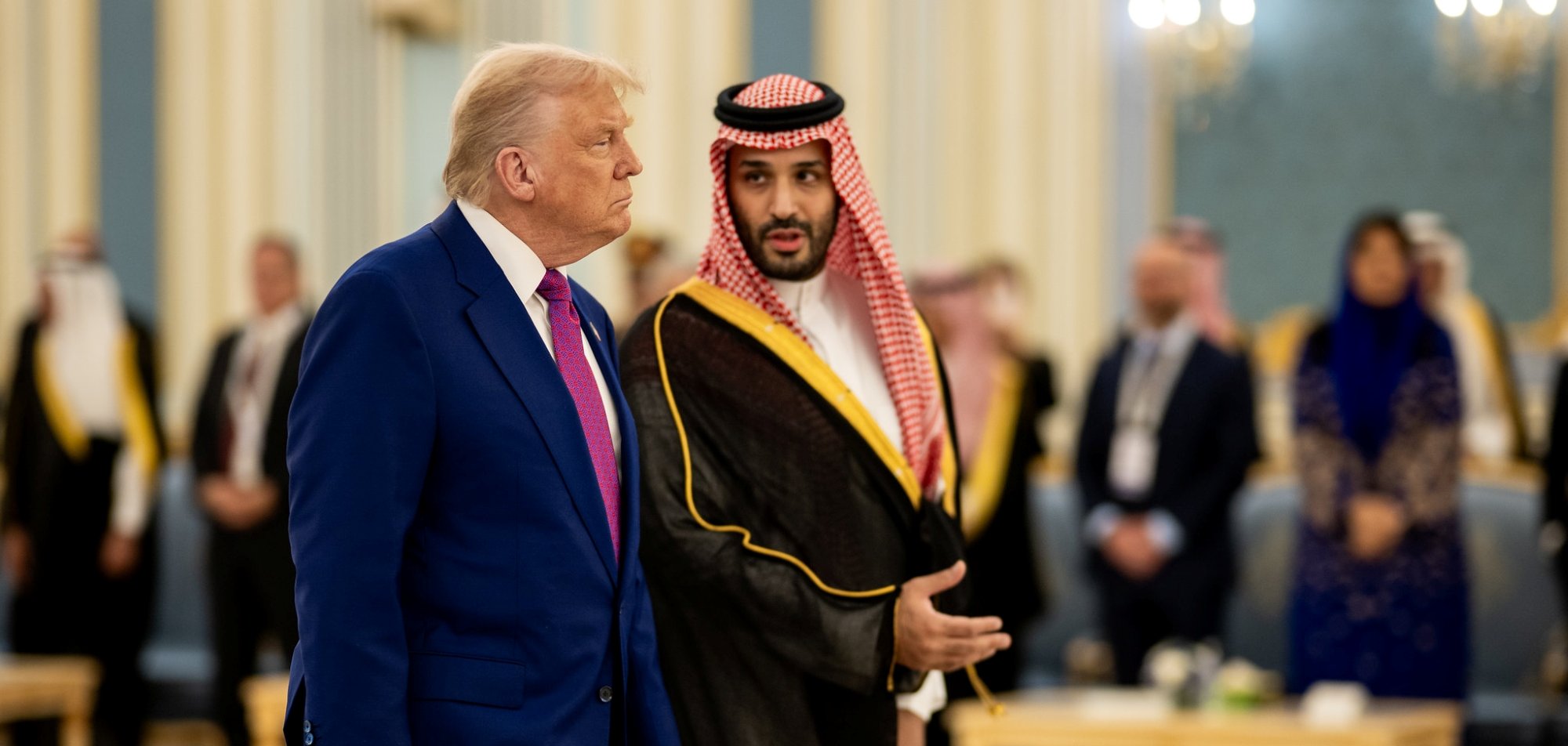 دو مرد در لباس‌های رسمی مختلف در یک محیط مجلل با نور ملایم و حضور افرادی دیگر.