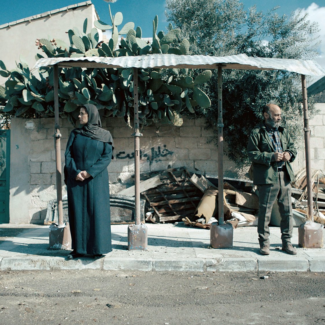 تظهر الصورة شخصين واقفين عند محطة انتظار. الشخص الأول امرأة ترتدي ملابس تقليدية وحجاب، وتقف بجانب جدار خلفي مزروع بأشجار الصبار. الشخص الثاني هو رجل يرتدي ملابس عسكرية ويبدو أنه مشغول بشيء في يده. في الخلفية، يمكن رؤية أجزاء من مبانٍ أخرى وبعض النفايات المتراكمة. السماء زرقاء، مما يدل على يوم مشمس. الجو العام يوحي بالهدوء والترقب.