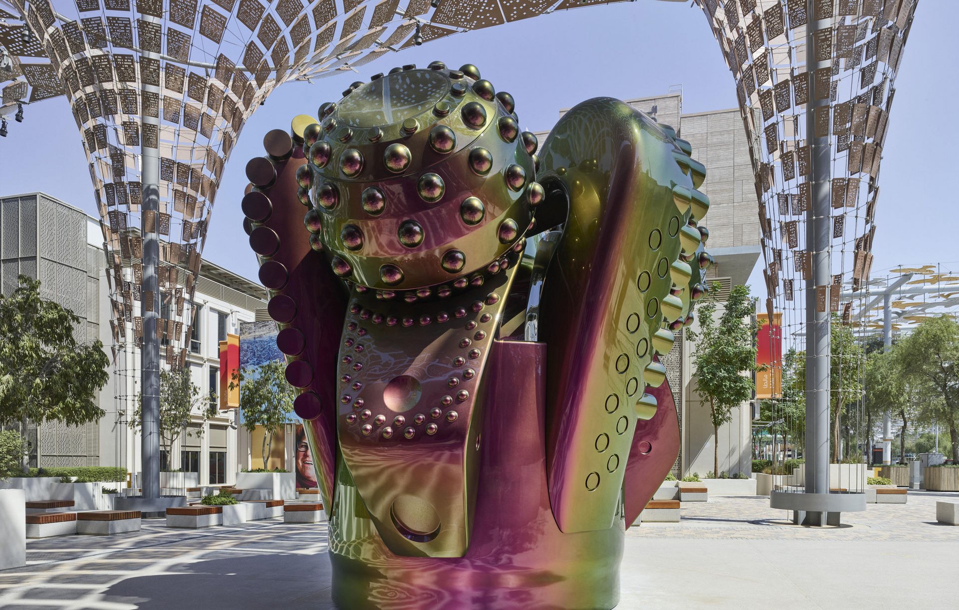 L'image présente une sculpture moderne au design futuriste, composée de formes organiques et géométriques. Les surfaces de la sculpture sont brillantes et colorées, avec des reflets irisés qui passent du vert au violet. Elle semble être placée sous une structure architecturale légère, qui offre de l'ombre et favorise une ambiance artistique. L'environnement autour de la sculpture est minimaliste, avec des arbres et quelques assises, ce qui met en valeur l'œuvre et crée une atmosphère apaisante.