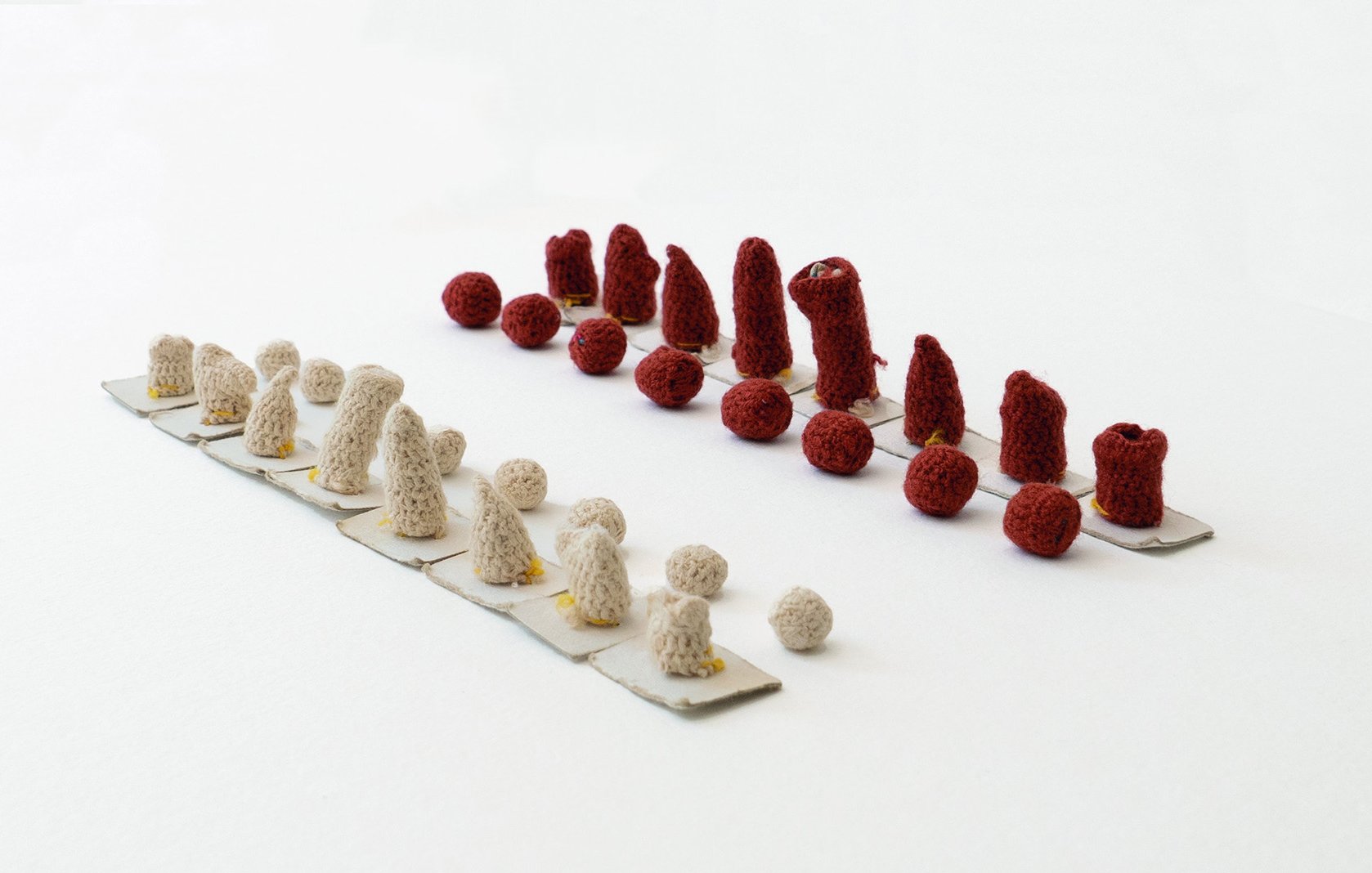 L'image montre une composition artistique composée de petits objets en crochet. Il y a deux groupes disposés sur une base claire : l'un en rouge et l'autre en beige. Les objets de chaque côté varient en forme et en taille, avec des cônes et des boules. La présentation est minimaliste, mettant en avant les textures et les couleurs des créations. L'arrangement est symétrique, créant un effet visuel intéressant.