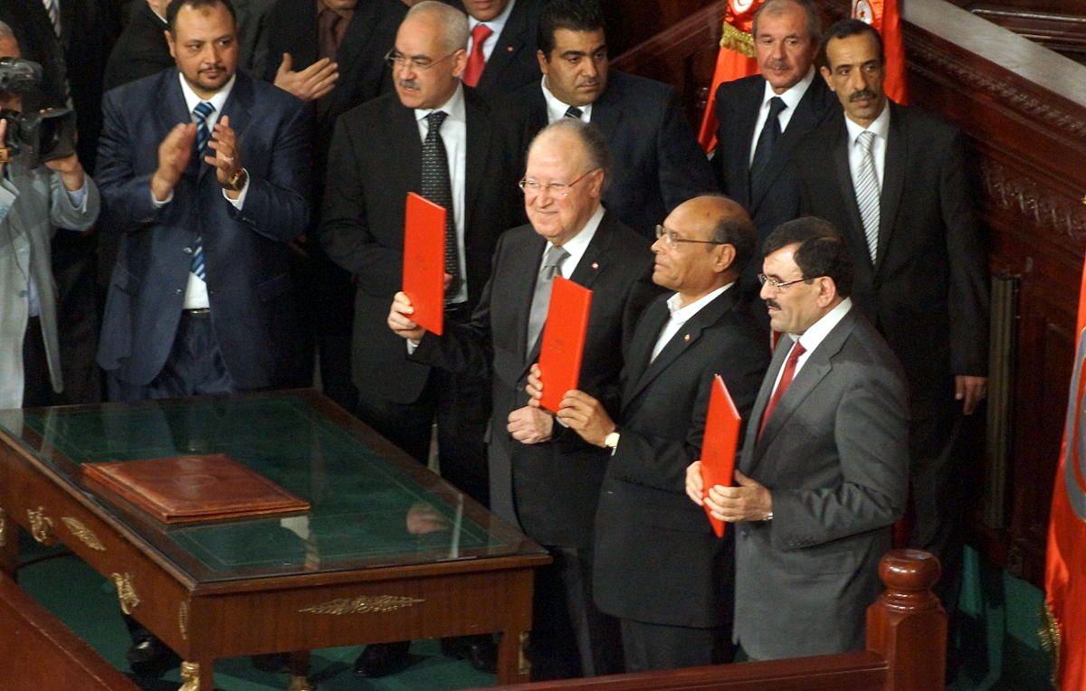 L'image montre un groupe d'hommes en costume, apparemment dans un cadre officiel, probablement lors d'une cérémonie ou d'un événement politique. Ils tiennent des documents en main, et l'ambiance semble solennelle. On peut voir une table en bois devant eux ainsi que des drapeaux en arrière-plan. Les personnes présentes semblent applaudir ou se concentrer sur l'événement.