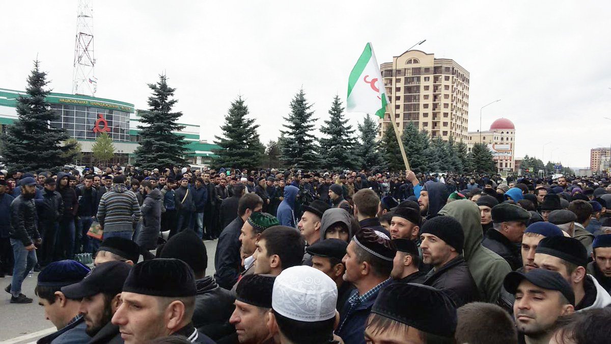 L'image montre une grande foule rassemblée dans une rue, probablement lors d'une manifestation ou d'un rassemblement public. Les participants semblent en majorité masculins et portent différents types de couvre-chefs. Certains tiennent des drapeaux, et l'atmosphère semble mobilisée. En arrière-plan, on peut voir des bâtiments et des arbres, ce qui donne un aperçu de l'environnement urbain. Le ciel est nuageux, ce qui pourrait indiquer un temps frais.