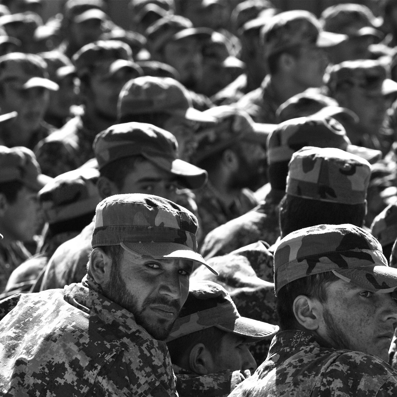 L'image montre un grand groupe de soldats en uniforme militaire, portant des casquettes assorties. Ils sont rassemblés et semblent attentifs, avec des expressions variées sur leurs visages. Le noir et blanc de la photo accentue les détails des uniformes camouflés et crée une atmosphère sérieuse, soulignant la discipline et la solidarité au sein du groupe.