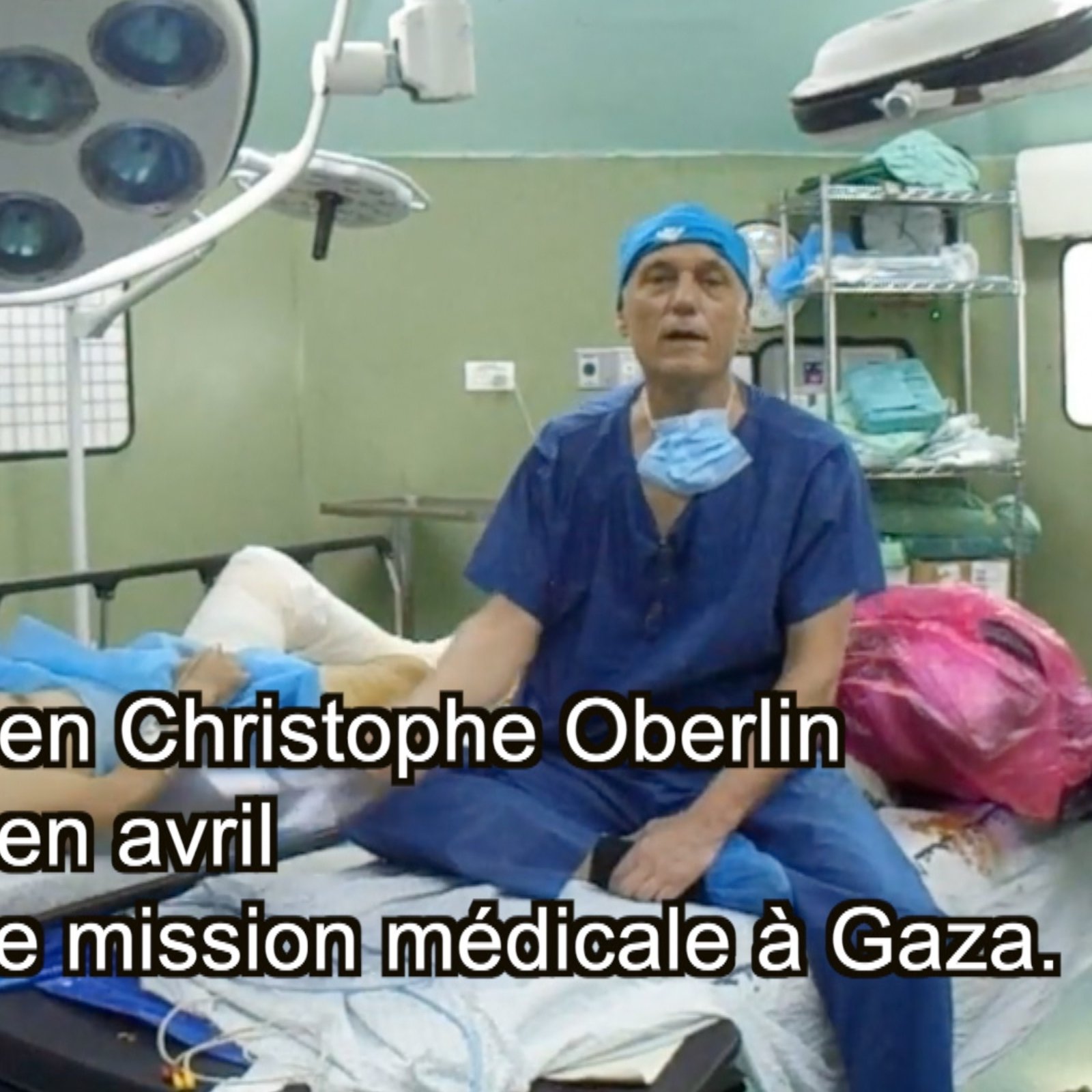L'image montre une scène dans une salle d'opération. On y voit plusieurs professionnels de la santé en train de travailler. Ils portent des blouses et des masques de protection caractéristiques des environnements médicaux. Au centre, un patient est couché sur une table d'opération, tandis qu'un chirurgien, probablement Christophe Oberlin, s'adresse à la caméra. Des équipements médicaux, tels que des lampes et des instruments chirurgicaux, sont visibles dans la pièce. Le texte indique que cette intervention a eu lieu en avril lors d'une mission médicale à Gaza.