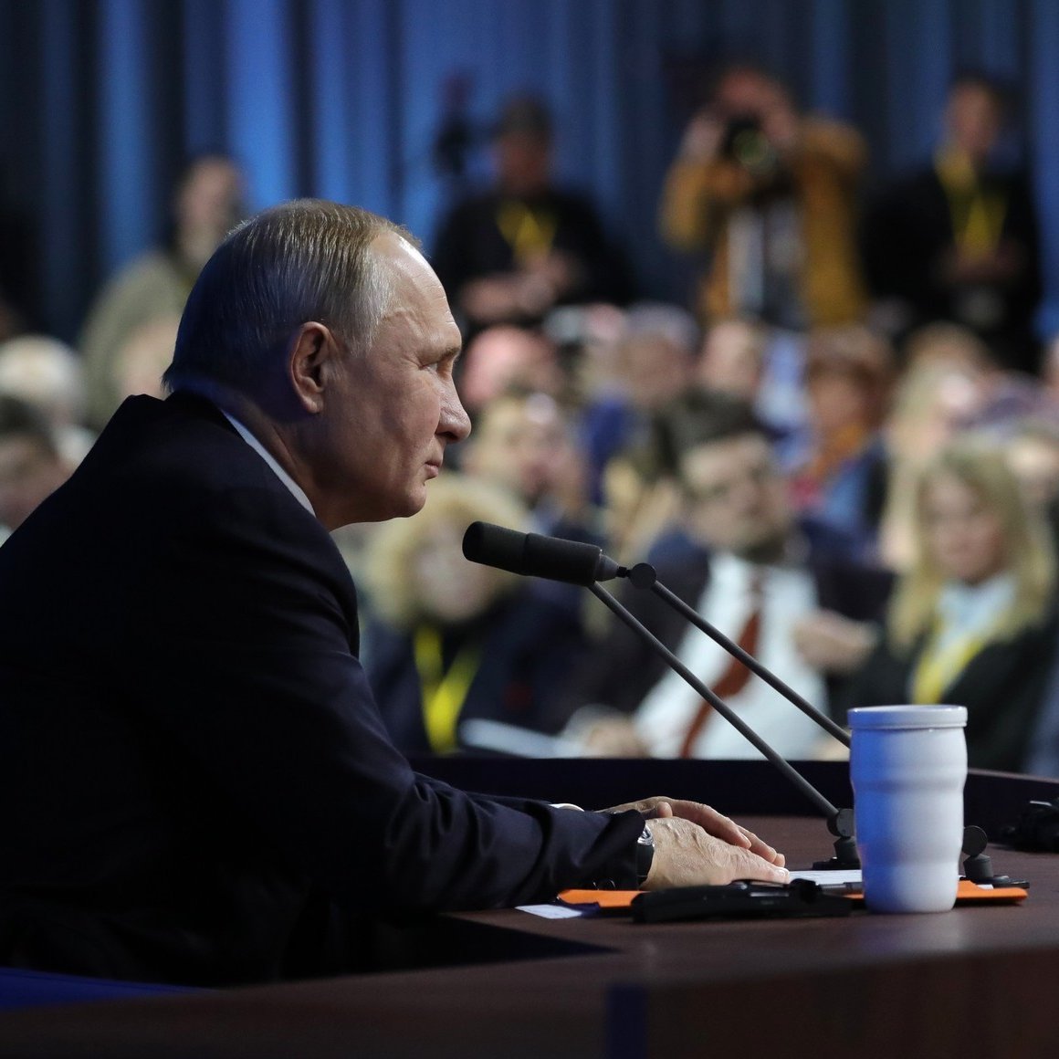 در این تصویر، ولادیمیر پوتین، رئیس‌جمهور روسیه، در حال سخنرانی یا پاسخ به سؤالات در یک کنفرانس مطبوعاتی به نظر می‌رسد. او در حال نشستن در کنار یک میز است که میکروفون دارد و در مقابل او تعدادی از خبرنگاران و حضار به نظر می‌رسند. محیط اطراف کمی تاریک‌تر است و تمرکز اصلی بر روی پوتین قرار دارد. تعدادی از افراد حاضر در جمع در پس‌زمینه دیده می‌شوند.