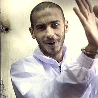 L'image montre un homme au regard confiant, portant une combinaison blanche. Ses mains sont menottées, et il semble faire un geste de la main, peut-être en souriant ou en se moquant de la situation. Le fond de l'image est flou, ce qui met l'accent sur l'homme. L'atmosphère générale donne une impression d'intensité, et l'homme semble détaché ou à l'aise malgré les menottes.