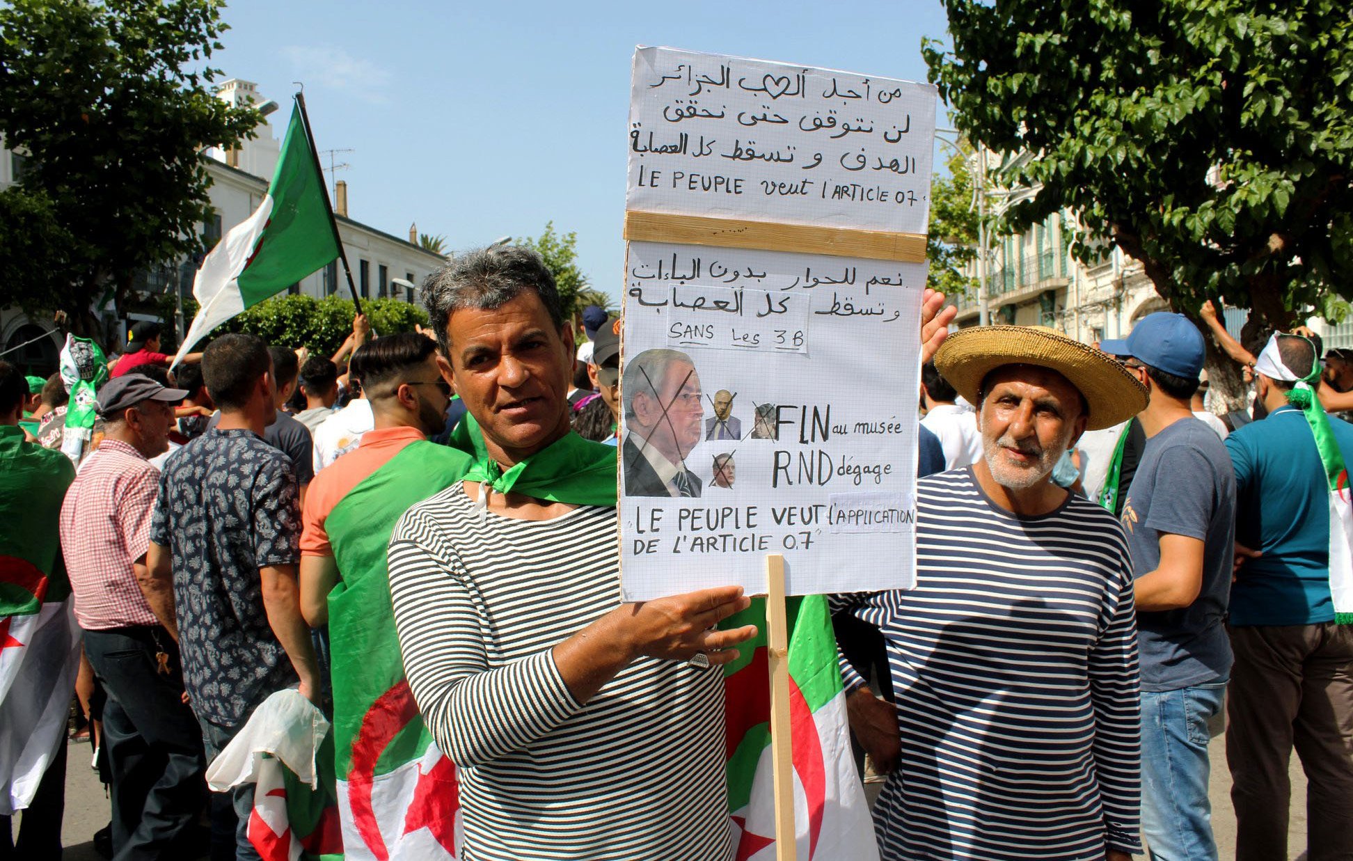 L'image montre un groupe de personnes manifestant, principalement des hommes, certains portant des drapeaux algériens. Deux manifestants se tiennent en avant-plan, l'un tenant une pancarte avec des inscriptions en arabe et en français. La pancarte semble exprimer des revendications politiques, notamment la fin de certaines pratiques liées à la politique algérienne. Les manifestants semblent unis et déterminés, se rassemblant pour faire entendre leur voix. L'atmosphère générale est celle d'une protestation pacifique.