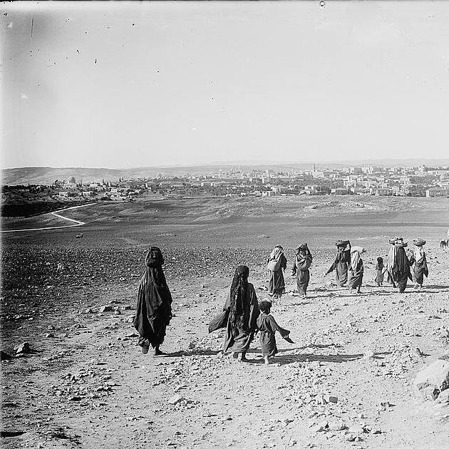 تصویر یک منظره تاریخی را نشان می‌دهد که در آن گروهی از افراد در حال پیاده‌روی در یک منطقه کوهستانی یا خشک هستند. آنها لباس‌های سنتی پوشیده‌اند و به نظر می‌رسد که به سوی یک روستا یا شهر در دوردست می‌روند. در پس‌زمینه، ساختمانی یا شهرهایی با زمینه کوهستانی مشخص است و جاده‌ای نیز در این تصویر دیده می‌شود. فضایی آرام و طبیعی حاکم است.