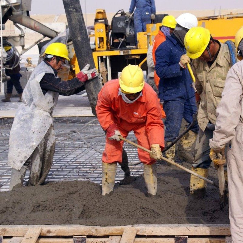 در این تصویر گروهی از کارگران مشغول به کار در یک پروژه ساختمانی هستند. آنها کلاه ایمنی به سر دارند و لباس‌های کار پوشیده‌اند. چند نفر در حال ریختن بتن و استفاده از تجهیزات مختلف هستند. محیط کار شلوغ و صنعتی به نظر می‌رسد و ماشین‌آلاتی در پس‌زمینه دیده می‌شود. کارگران با جدیت و همکاری در حال انجام وظایف خود هستند.