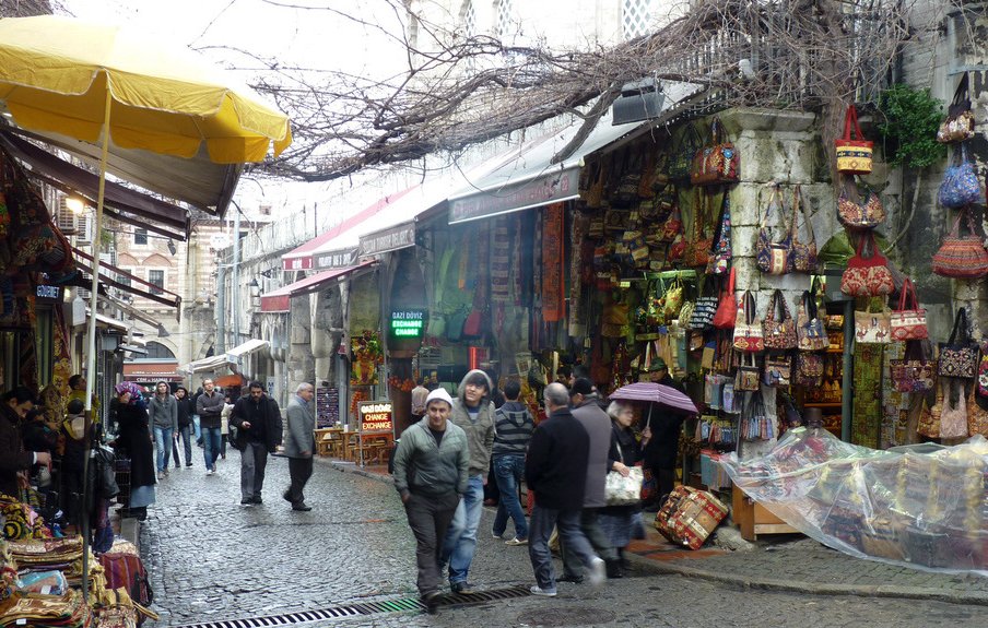 L'image montre une ruelle animée d'un marché. Les pavés sont humides, suggérant peut-être qu'il a récemment plu. Les étals sont remplis de sacs colorés et d'autres articles artisanaux, donnant une impression de vivacité et de diversité. Des gens se déplacent dans la rue, certains se protégeant sous des parapluies. Au-dessus, des branches nues d'arbres créent une atmosphère presque nostalgique, tandis que des auvents jaunes ajoutent une touche de couleur à la scène.