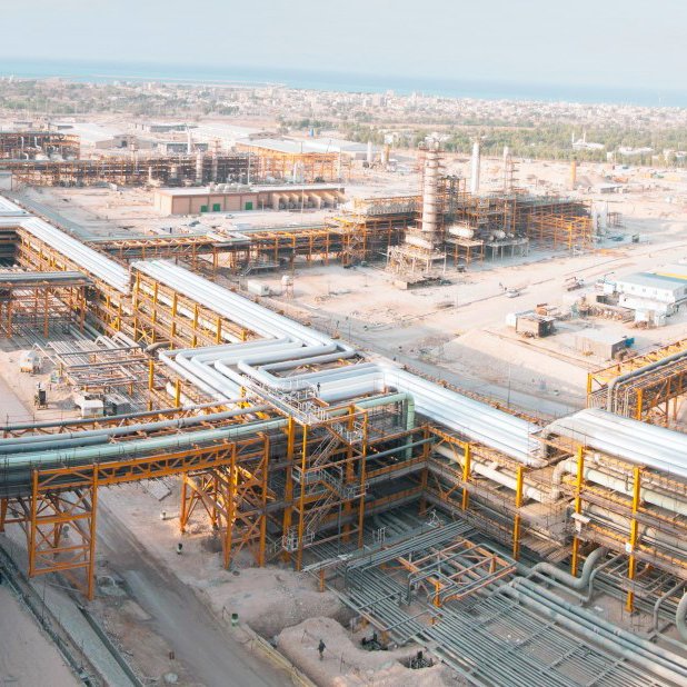 L'image montre un vaste complexe industriel, probablement lié à l'extraction ou au traitement des ressources naturelles. On peut voir de nombreuses structures métalliques, des tubes et des pipelines s'étendant sur une grande surface. L'environnement semble désertique ou aride, et au loin, on aperçoit la mer. La disposition des installations indique un environnement de travail organisé, avec des infrastructures complexes qui témoignent d'une activité industrielle importante.