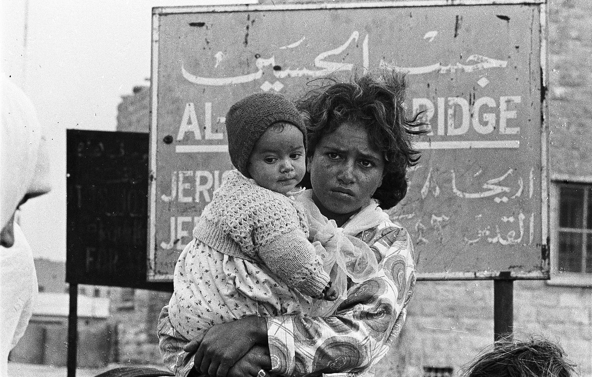 The image depicts a young woman holding a baby, both appearing to be in a challenging environment. The woman has a serious expression on her face, and her hair is unkempt. The baby, swaddled in a heavy garment, is held close to her. In the background, there's a sign written in Arabic, which seems to indicate a location, possibly related to a city or area. The overall atmosphere conveys a sense of hardship and resilience. The image is in black and white, emphasizing the emotional weight of the scene.