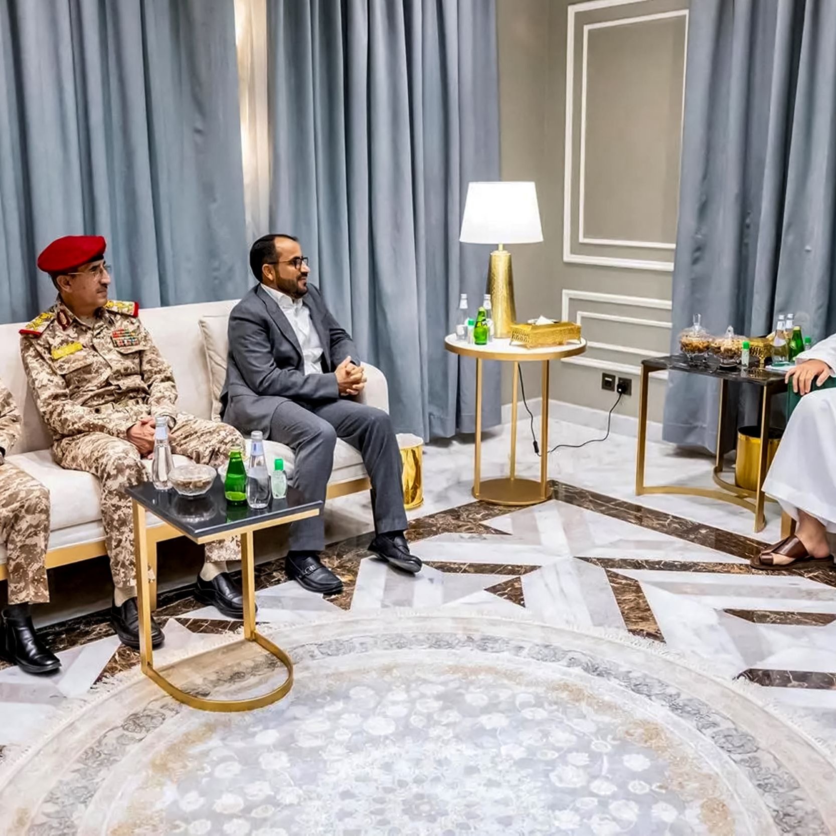 تظهر الصورة مجموعة من الأشخاص الذين يجلسون في غرفة معيشة أنيقة. في الجهة اليسرى، يوجد ضابطان عسكريان يرتديان زيًا عسكريًا، أحدهما يرتدي قبعة حمراء. بينما يجلس رجل ثالث في المنتصف بزي مدني. على اليمين، يوجد رجل يرتدي لباسًا تقليديًا سعوديًا (ثوب وغطاء رأس) يجلس على أريكة. يوجد أيضًا طاولة صغيرة أمامهم تحتوي على بعض المشروبات. الجدران مكسوة بستائر تتناسب مع الديكور الداخلي.