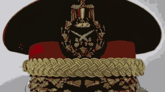 L'image montre un uniforme militaire, plus précisément un couvre-chef. Il s'agit d'une casquette ou d'un képi avec une visière. Le chapeau est décoré de divers ornements en métal ou en broderie, incluant un insigne central représentant des attributs militaires. On peut observer également une bordure en or ou en couleur claire qui donne une impression de prestige. Les détails suggèrent que ce couvre-chef est associé à un grade élevé dans une force armée.