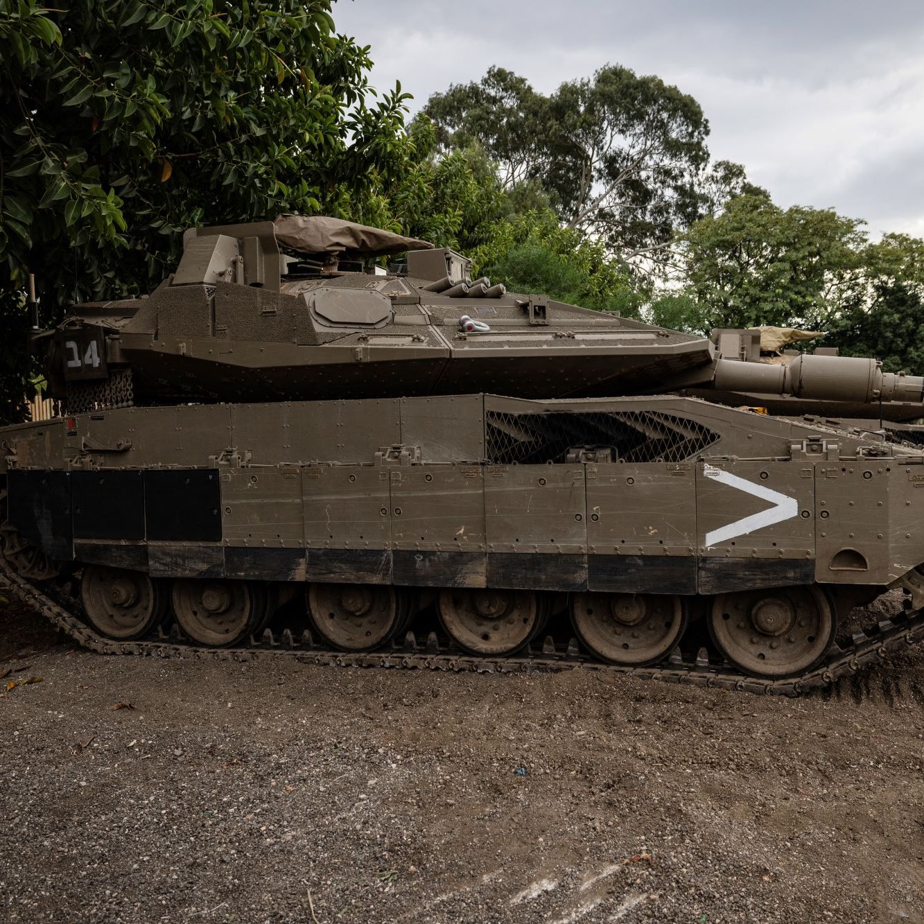 الصورة تظهر دبابة مدرعة على الأرض، متوقفة بجانب مجموعة من الأشجار. الدبابة تحمل لوناً زيتونياً وتبدو قوية ومتينة، مع تفاصيل واضحة مثل البرج والمدفع. السماء خلفها ملبدة بالغيوم، مما يعطي انطباعًا عن أجواء درامية. الإطار العام يوحي بمكان عسكري أو منطقة تدريب.