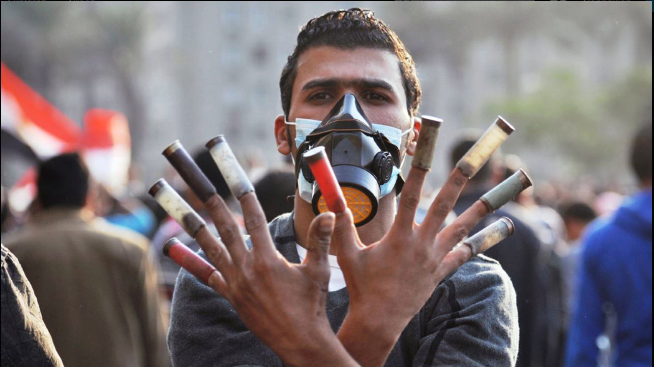 الصورة تظهر شابًا يرتدي كمامة واقية بينما يرفع يديه للتعبير عن موقفه. تظهر أصابعه تحمل أنابيب ملونة، مما قد يشير إلى استخدامه لأدوات معينة في سياق احتجاج أو تجمع شعبي. الخلفية تبدو غير واضحة ولكنها تشير إلى وجود حشد من الناس، مما يدل على أن الصورة قد التقطت في مكان يشهد تظاهرات أو فعاليات سياسية. تعبر الصورة عن مشاعر قوية من المقاومة والتحدي.