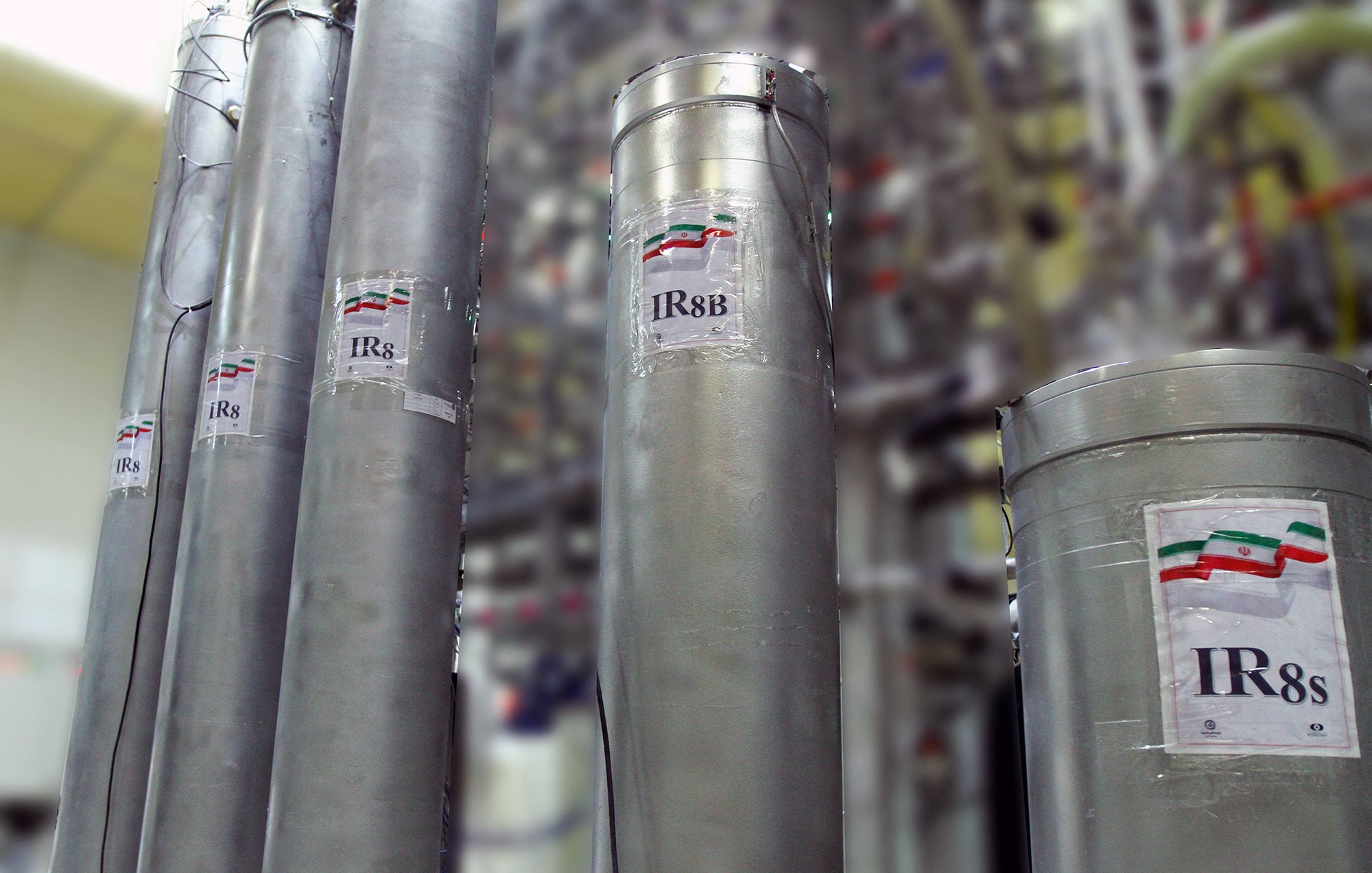 تظهر في الصورة مجموعة من الأسطوانات المعدنية، وكل منها يحمل ملصق مكتوب عليه "IR8" أو "IR8s" مع رمز علم دولة إيران. خلف هذه الأسطوانات، يمكن رؤية مجموعة من الأنابيب والتجهيزات الأخرى، مما يشير إلى أنها قد تكون جزءًا من منشأة صناعية أو مركز بحث علمي. الجو العام في الصورة يعكس بيئة تقنية عالية ومجهزة بأجهزة متطورة.