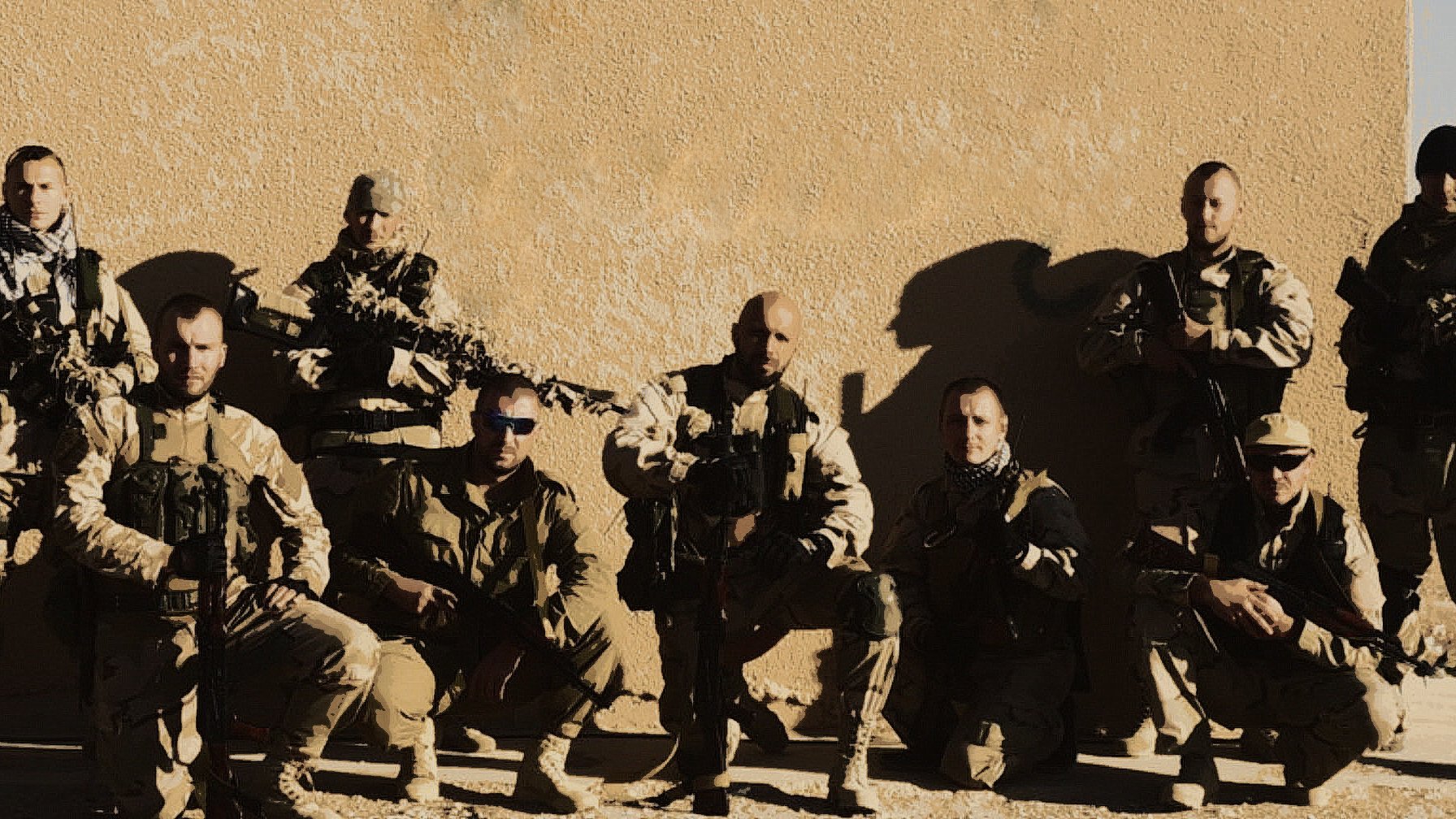 The image appears to show a group of military personnel posing together against a neutral-colored background. They are dressed in tactical gear, including vests and uniforms, and are equipped with firearms. The group is arranged in two rows, with some members kneeling and others standing. The overall tone of the image suggests a camaraderie among the team. The lighting emphasizes their serious expressions and the rugged environment.