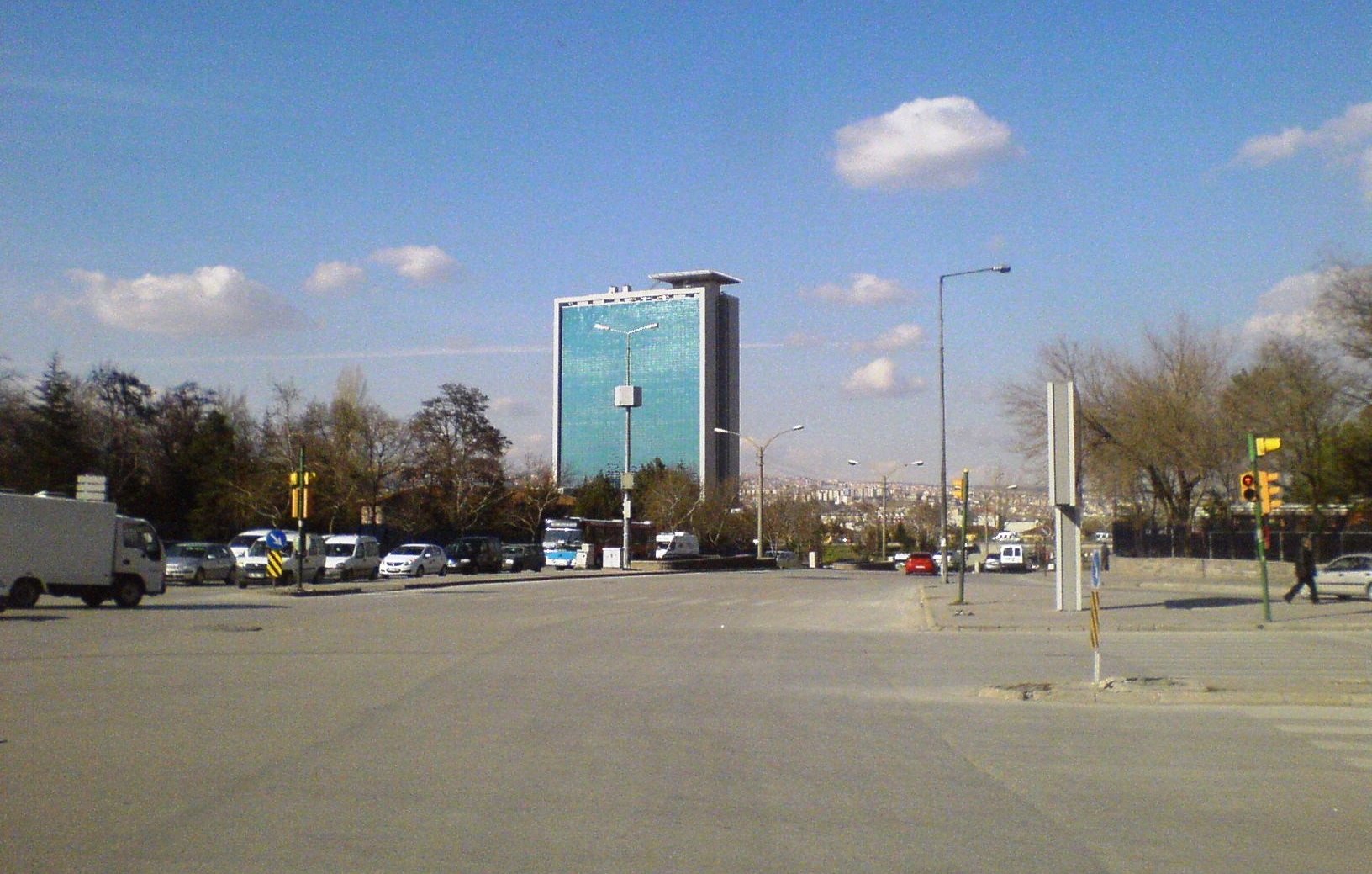 L'image montre une vue d'une rue avec un bâtiment moderne en arrière-plan. Ce bâtiment, de grande hauteur, a une façade en verre qui reflète le ciel. Le ciel est clair avec quelques nuages dispersés. On peut voir des voitures garées et circulant sur la route, ainsi que des arbres sur le côté. L'ensemble donne une impression de ville moderne et animée.