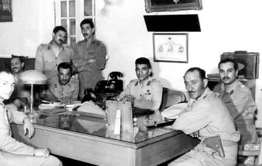 L'image montre un groupe de six hommes en uniforme militaire réunis autour d'une grande table en bois dans un bureau. Certains d'entre eux sont assis, tandis que d'autres se tiennent derrière. L'environnement décoré de manière classique inclut des éléments tels qu'une lampe de bureau et des tableaux accrochés au mur. L'atmosphère semble formelle, et les expressions des hommes varient entre concentration et sérieux.