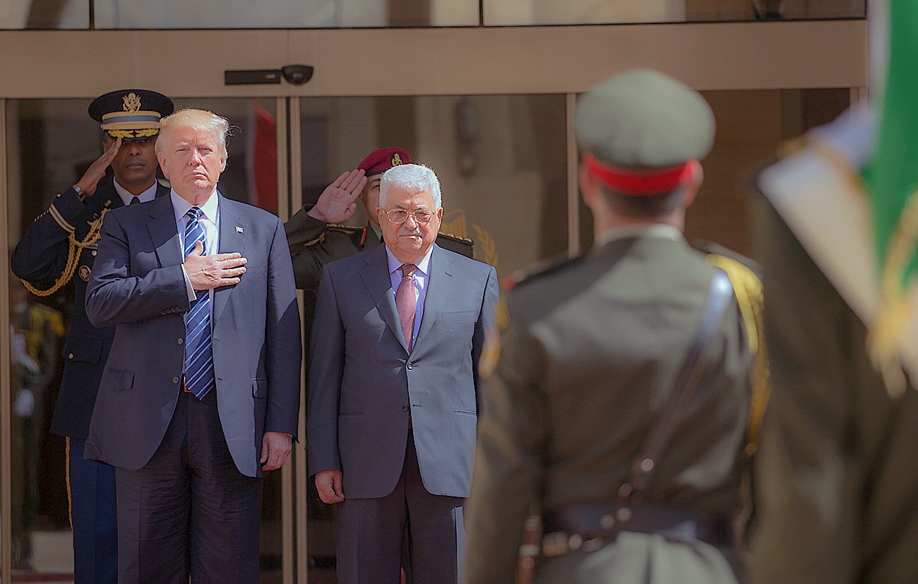 در تصویر، دو نفر در حال ایستادن هستند. یکی از آن‌ها در سمت چپ، در یک لباس رسمی به رنگ تیره و با کراواتی آبی، دست خود را روی قلبش گذاشته و به جلو نگاه می‌کند. نفر دیگر در سمت راست، یک کت و شلوار خاکستری به تن دارد و به آرامی در کنار شخص اول ایستاده است. در پس‌زمینه، تعدادی از افراد نظامی ایستاده‌اند و نسبت به این دو نفر احترام می‌گذارند. فضای تصویر نشان‌دهنده یک مراسم رسمی است.