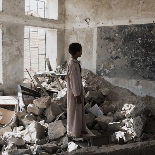 در این تصویر، یک پسر جوان در یک کلاس درس ویران شده ایستاده است. دیوارها و سقف کلاس آسیب دید‌ه‌اند و بخش‌هایی از زیرساخت‌ها به طور کامل تخریب شده‌اند. در اطراف او، زباله‌ها و آوارهای ساختمانی وجود دارد. یک تابلو سیاه با نوشته‌هایی بر روی دیوار وجود دارد، اما وضعیت آن نیز نامناسب است. نور از پنجره‌های شکسته به فضای داخلی می‌تابد و حس ناامیدی و ویرانی را منتقل می‌کند.