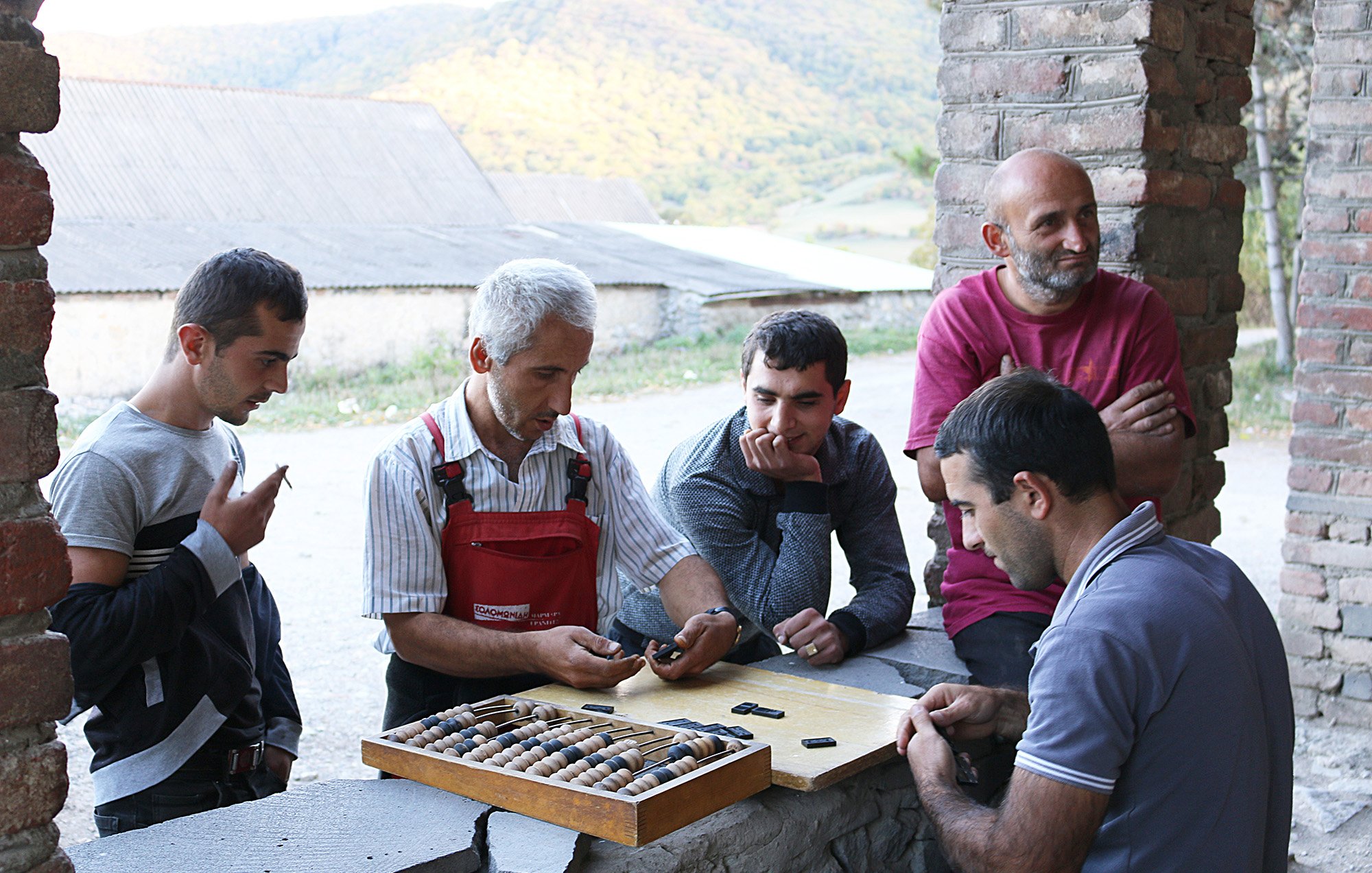 تصویر گروهی از مردان را نشان می‌دهد که در یک محیط روستایی گرد هم آمده‌اند. در مرکز تصویر، مردی با موهای سفید و لباس‌های کارگاهی به نظر می‌رسد که مشغول توضیح یا نشان دادن چیزی به دیگران است. مردان دیگر به دقت به او گوش می‌دهند و در حال بررسی یک صفحه بازی هستند که تعدادی مهره در آن قرار دارد. پس‌زمینه تصویر شامل تپه‌های سرسبز و یک ساختمان قدیمی است. جو عمومی تصویر دوستانه و غیررسمی به نظر می‌رسد.