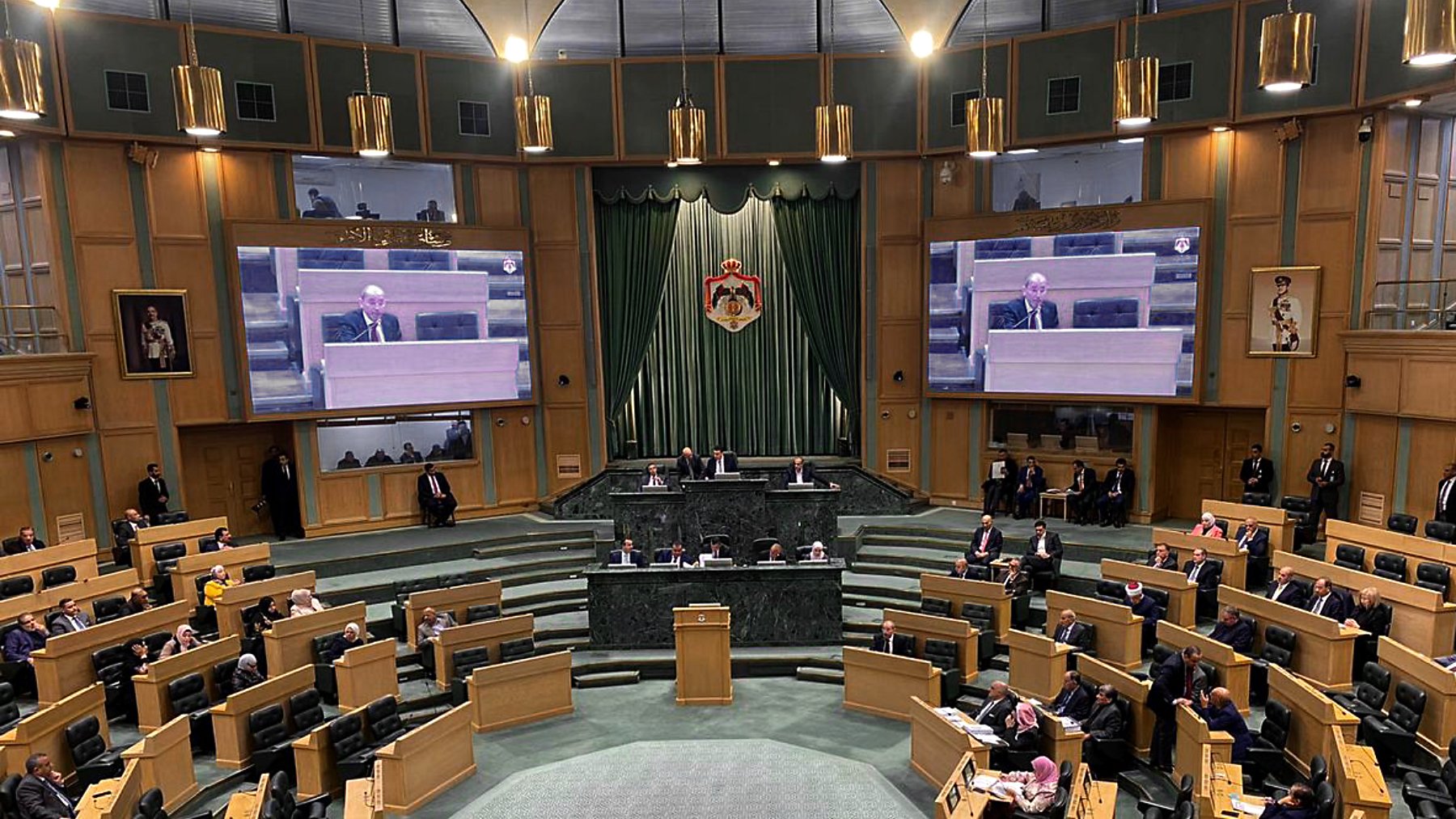 L'image montre l'intérieur d'une grande salle de réunion ou d'assemblée, probablement un parlement ou une assemblée législative. Au centre, une estrade est prévue pour les orateurs, et des écrans sont visibles sur les murs pour diffuser les interventions. On peut voir plusieurs membres assis sur des rangées en pente, organisés en sections. Les couleurs dominantes sont des tons de vert et de bois, créant une ambiance formelle. Des personnes sont également présentes debout, écoutant attentivement.