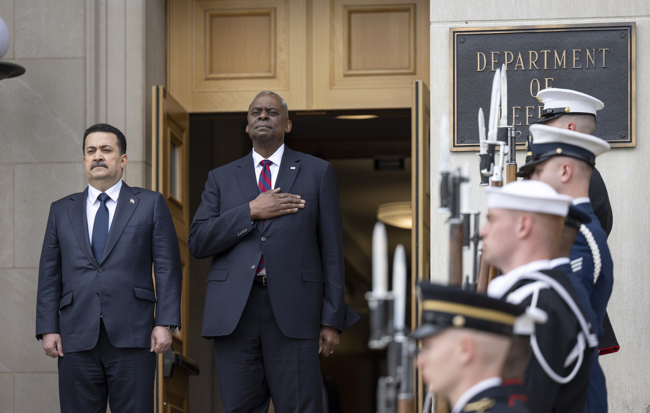 L'immagine mostra due uomini in piedi davanti all'entrata di un edificio governativo, probabilmente il Dipartimento della Difesa. Uno di loro indossa un abbigliamento formale con un completo scuro e una cravatta, mentre l'altro ha il braccio destro sul cuore, indicando un gesto di rispetto o di onore. Dietro di loro, ci sono diversi membri delle forze armate in uniforme, schierati con armi in mano, pronti a rendere onore. L'atmosfera è solenne e pomposa, suggerendo un evento ufficiale o una cerimonia.