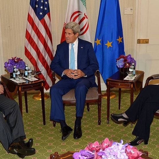 L'image montre une réunion en intérieur entre trois personnes assises dans un environnement élégant. À gauche, on voit un homme en costume noir, probablement un représentant de l'Iran. Au centre, un homme en costume clair, probablement un diplomate américain, semble discuter. À droite, une femme en blouse bleue, possiblement une diplomate européenne, écoute attentivement. Les participants sont entourés de tables avec des bouteilles d'eau et des fleurs en centre de table. L'atmosphère semble être celle de discussions diplomatiques sérieuses.