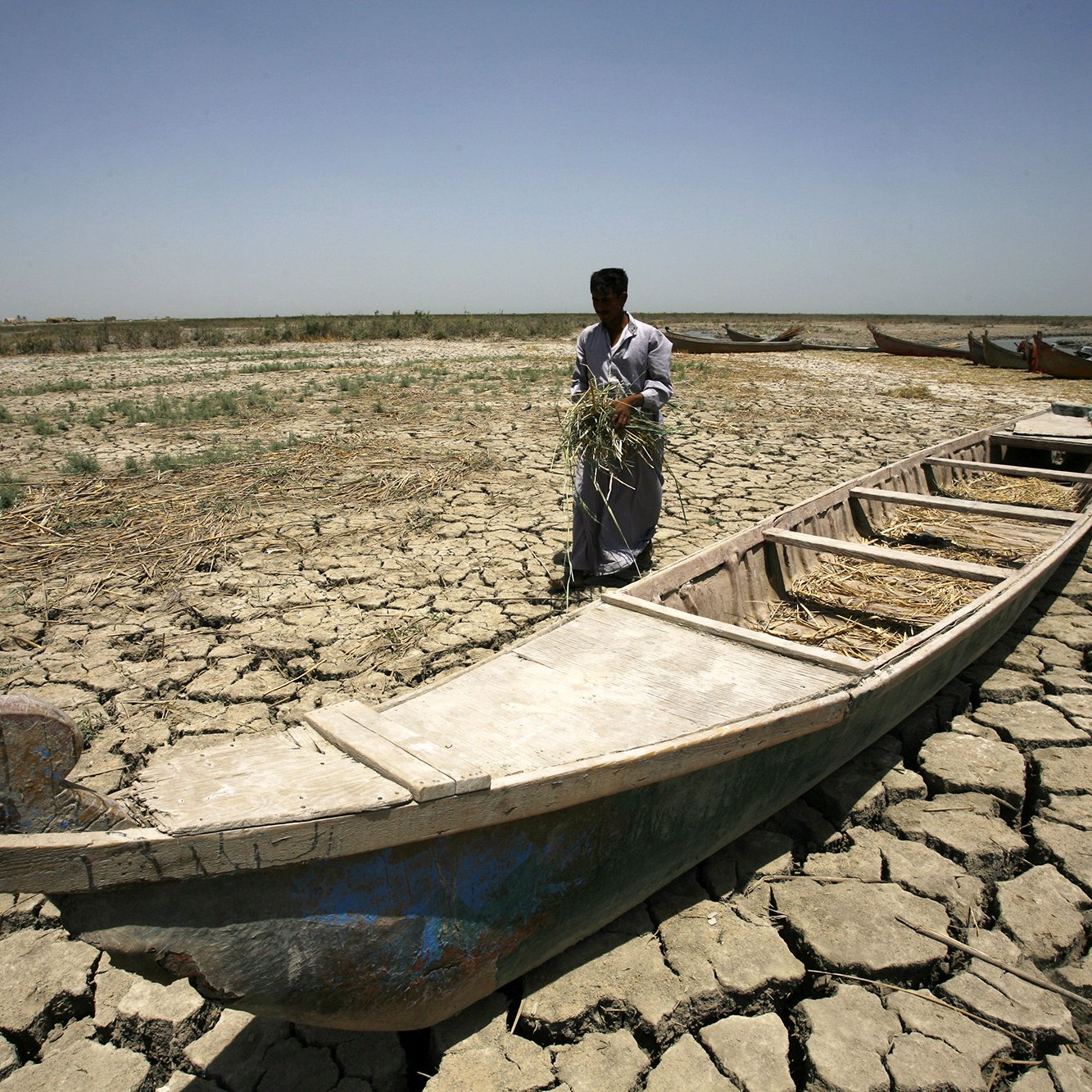تظهر في الصورة مشهداً لرجلاً يقف على أرض قاحلة ومجففة. هو يحمل بعض الأعشاب أو الحشائش، ويبدو أنه يقف بجانب قارب قديم مهجور. الأرض مكسوة بالشقوق، مما يشير إلى جفاف المنطقة. السماء صافية والشمس ساطعة، مما يعطي انطباعاً عن مناخ جاف وصعب. القوارب الأخرى تظهر في الخلفية، لكنها أيضاً تبدو غير مستخدمة.