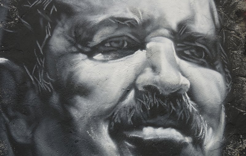L'image représente une œuvre murale en noir et blanc, mettant en avant un visage masculin avec des traits expressifs. La personne a une moustache proéminente, et son expression semble montrer une certaine intensité. Les détails des ombres et des lumières apportent de la profondeur au portrait, accentuant les émotions qui s'en dégagent. Les contours sont soigneusement travaillés, ce qui donne une apparence réaliste au personnage.