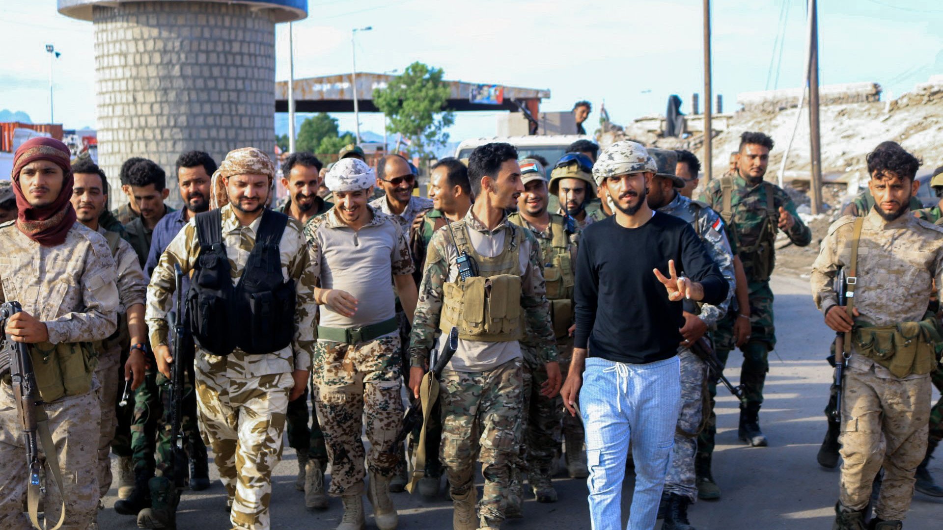 The image shows a group of soldiers and military personnel walking together in an outdoor setting. The soldiers are wearing various military uniforms and gear, including vests and helmets. Some are carrying weapons. In the foreground, a man in civilian clothes, showing a peace sign with his fingers, stands among them, smiling. The background suggests an urban area, with buildings and a road visible. The overall atmosphere appears to be one of camaraderie and unity.