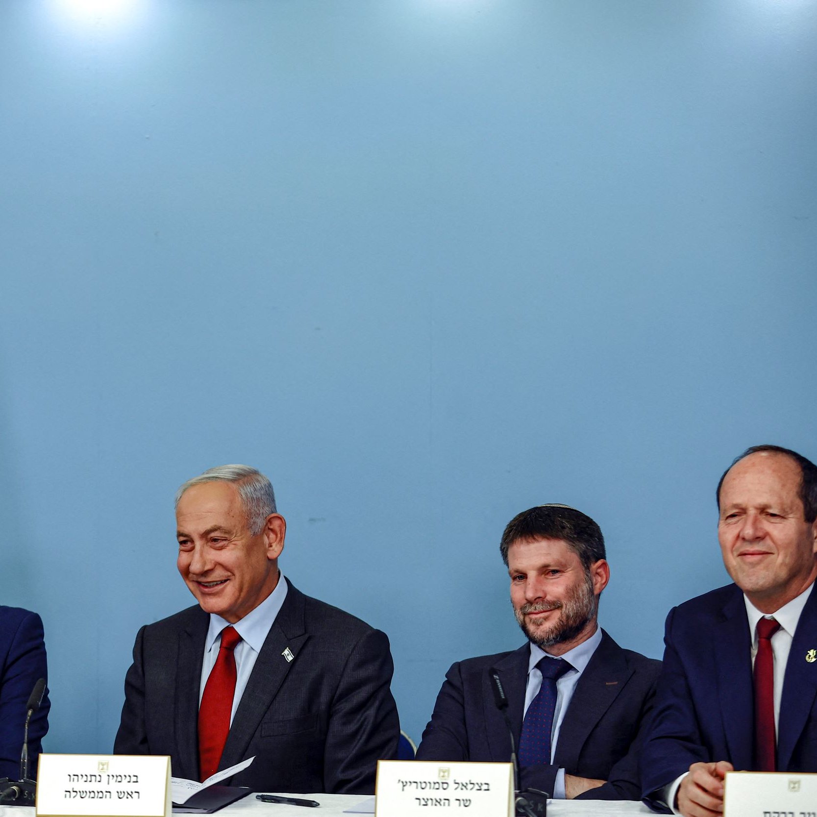 L'image montre un groupe de quatre hommes assis à une table, souriant et discutant. En arrière-plan, il y a un drapeau israélien. Les hommes portent des costumes et des cravates, et il semble qu'ils soient en train de participer à une conférence de presse ou à un événement officiel. L'ambiance est décontractée mais professionnelle.