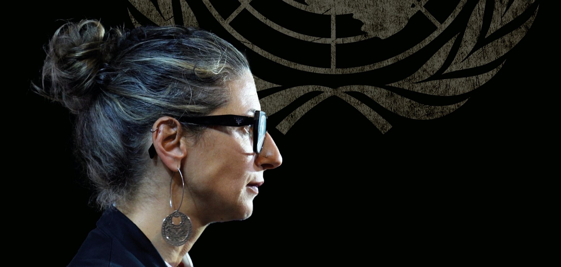 Donna con occhiali e capelli raccolti, in un ambiente con il logo delle Nazioni Unite sullo sfondo.