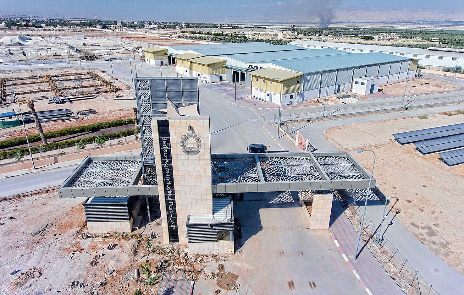 تظهر الصورة مشهداً جوياً لمدخل منشأة أو منطقة صناعية. يتوسط المدخل بناء حديث ذو تصميم معماري مميز، مع واجهة تحتوي على عناصر فنية تتضمن الزخارف والأشكال الهندسية. خلف المدخل، تمتد المستودعات والمبانِ الصناعية، مع تواجد أرضيات مرصوفة ومناطق خضراء ذات أشجار. في الأفق، يمكن رؤية دخان يخرج من أحد المواقع، مما يشير إلى نشاط صناعي، بينما يتواجد في الجانب الأيسر ألواح شمسية. الجو العام يبدو هادئاً ومنظمًا، مع مساحات شاسعة من الأرض.