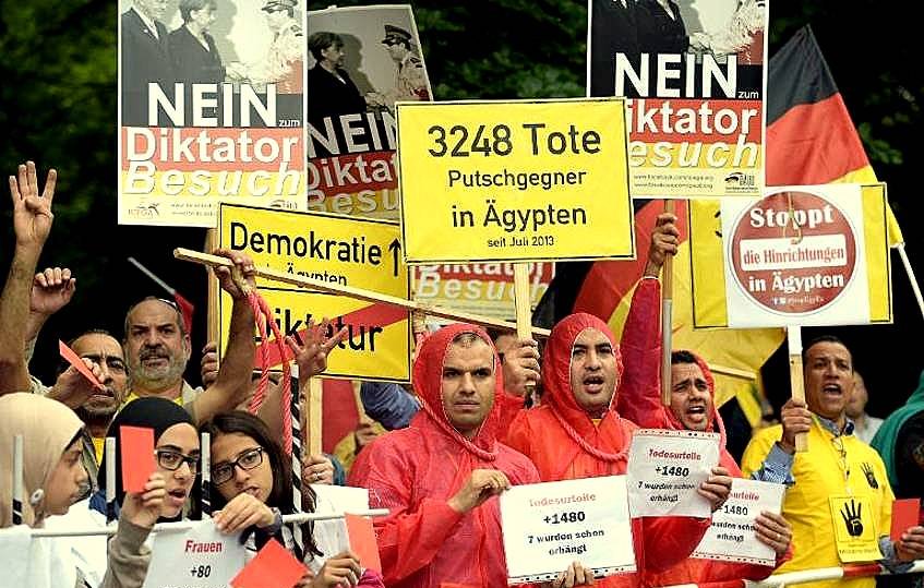 L'image montre un groupe de manifestants rassemblés pour une protestation. Ils portent des vêtements de pluie rouges et tiennent des pancartes avec des messages en allemand et en anglais, dénonçant une dictature et parlant de décès d'opposants en Égypte. On peut voir des slogans comme "NEIN" et des chiffres indiquant le nombre de morts. L'atmosphère semble sérieuse, témoignant de l'engagement des manifestants pour leurs causes.