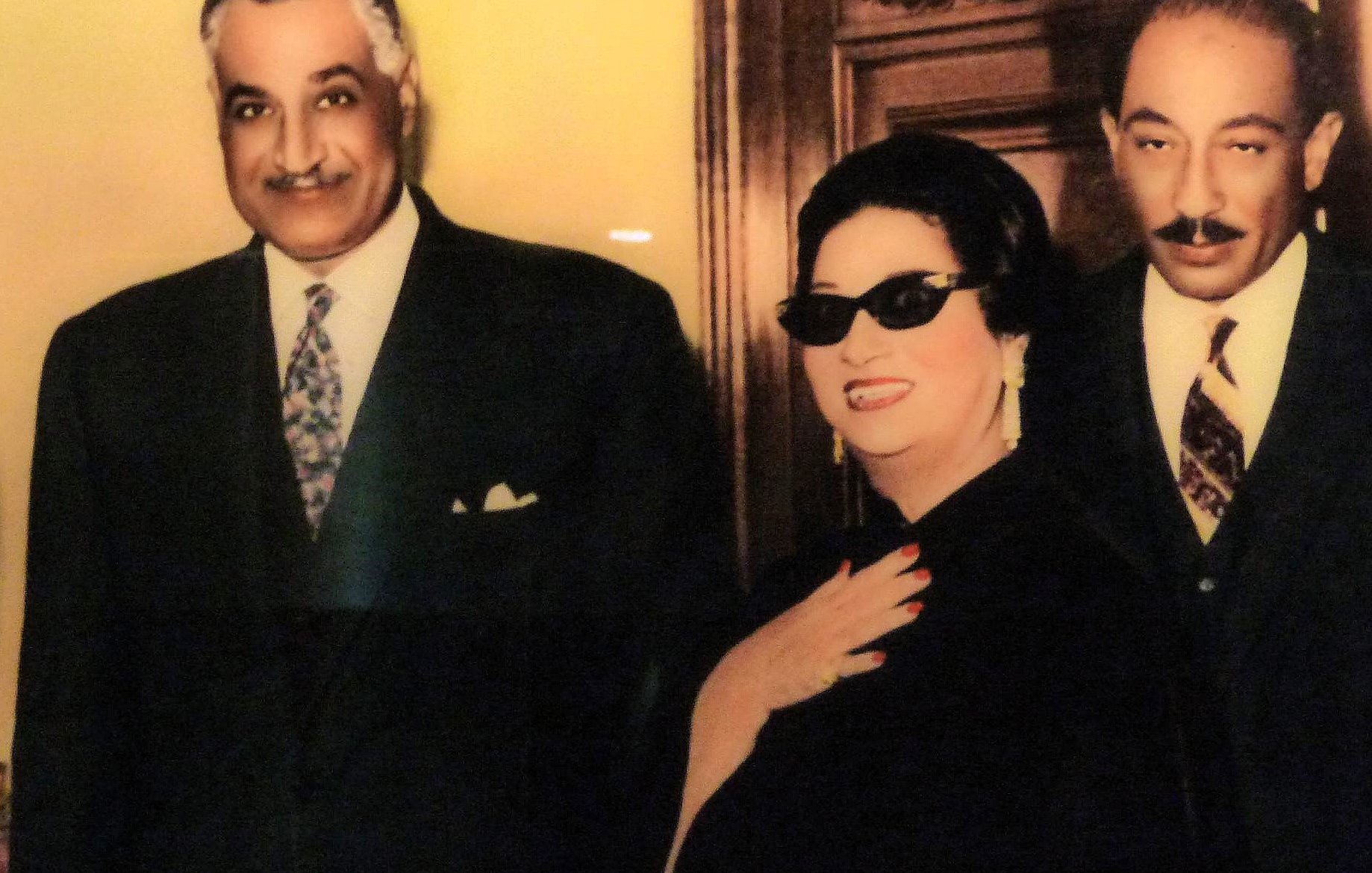 L'image montre trois personnes bien habillées posant ensemble. À gauche, un homme souriant porte un costume avec une cravate. Au centre, une femme en robes sombres et lunettes de soleil affiche un sourire radieux et une pose élégante. À droite, un autre homme en costume, qui semble sérieux, se tient près d'elle. L'arrière-plan est décoré de manière raffinée, mettant en valeur l'ambiance formelle de la photo.