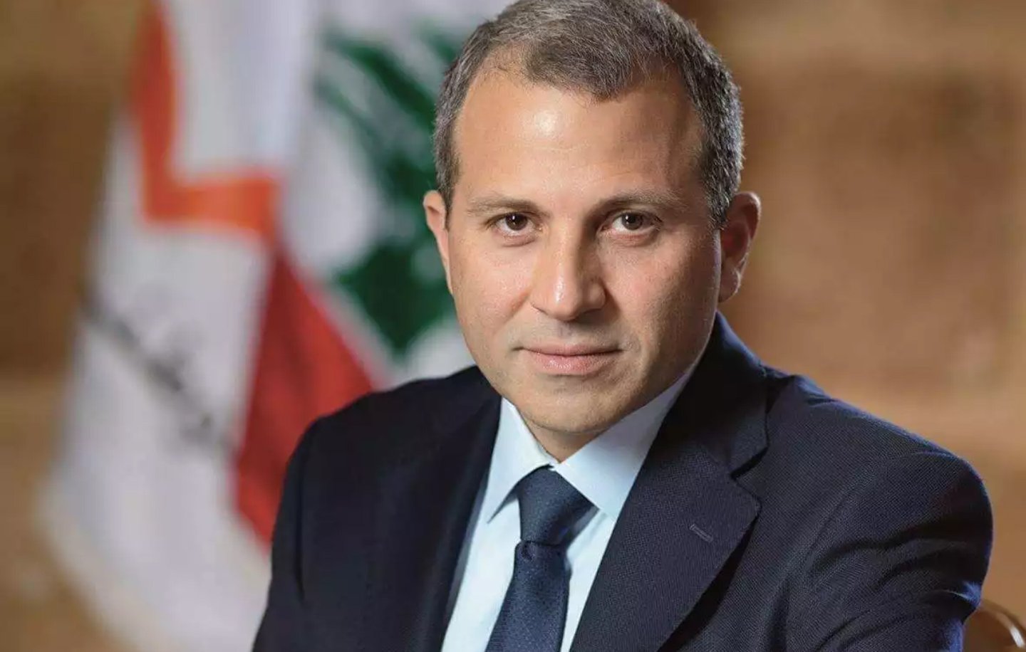 الصورة تظهر رجلًا يرتدي بدلة رسمية داكنة، مع قميص أبيض وروابط عنق زرقاء. يجلس بشكل مشي هادئ وملامحه تظهر الثقة. في الخلفية، تتواجد علم لبنان، مما يشير إلى احتمال ارتباطه بالسياسة أو الحكومة في لبنان. الجو العام للصورة يعكس الرسمية والجدية.