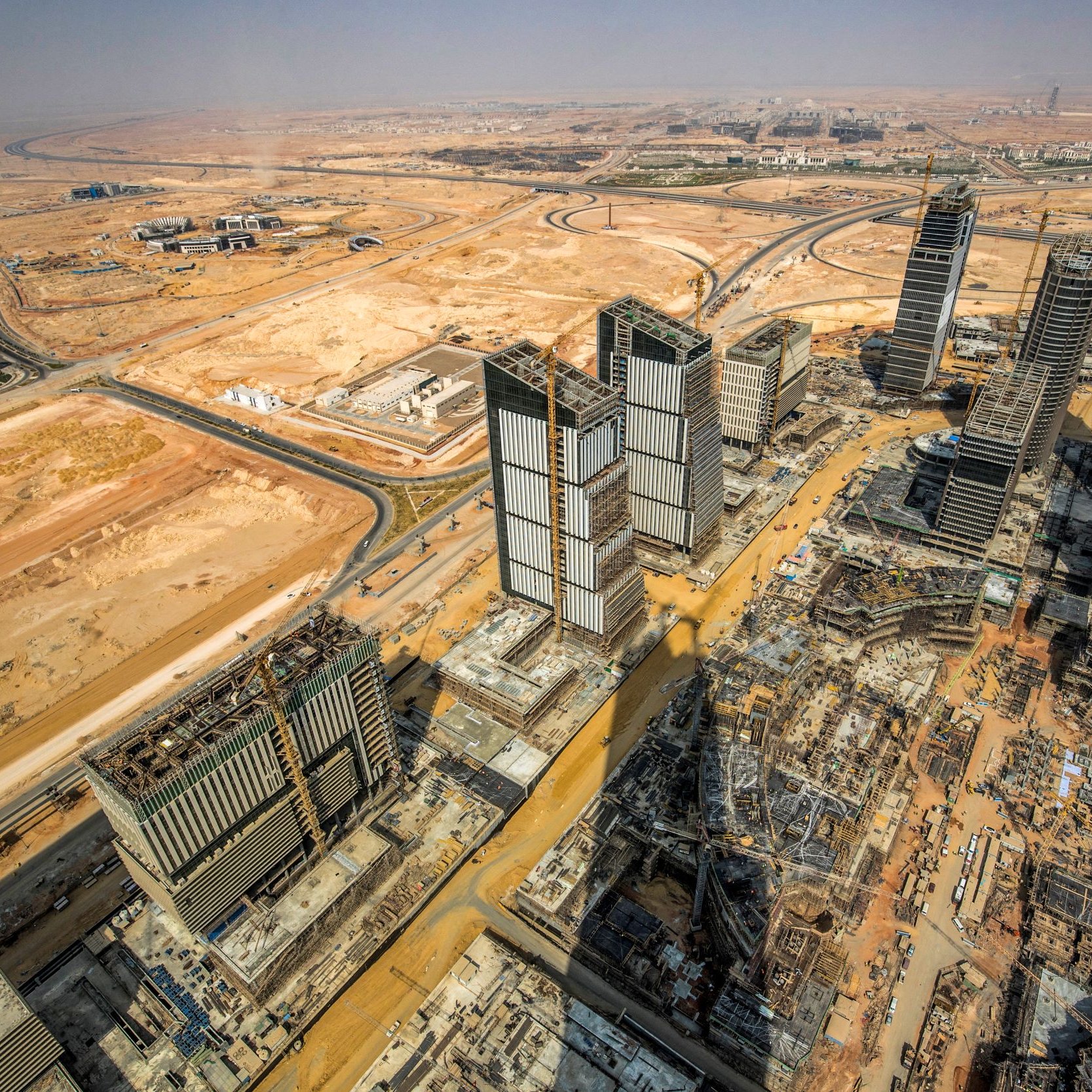 تظهر الصورة منظرًا جويًا لمدينة قيد الإنشاء في منطقة صحراوية. يمكن ملاحظة العديد من الأبراج والمباني الحديثة التي تُشيد، بالإضافة إلى الطرق الواسعة التي تلتف حول المشروع. يحيط بالمكان أرض جرداء، مما يبرز التباين بين البناء الحديث والطبيعة القاحلة. كما تُظهر الصورة التطور العمراني السريع في تلك المنطقة.