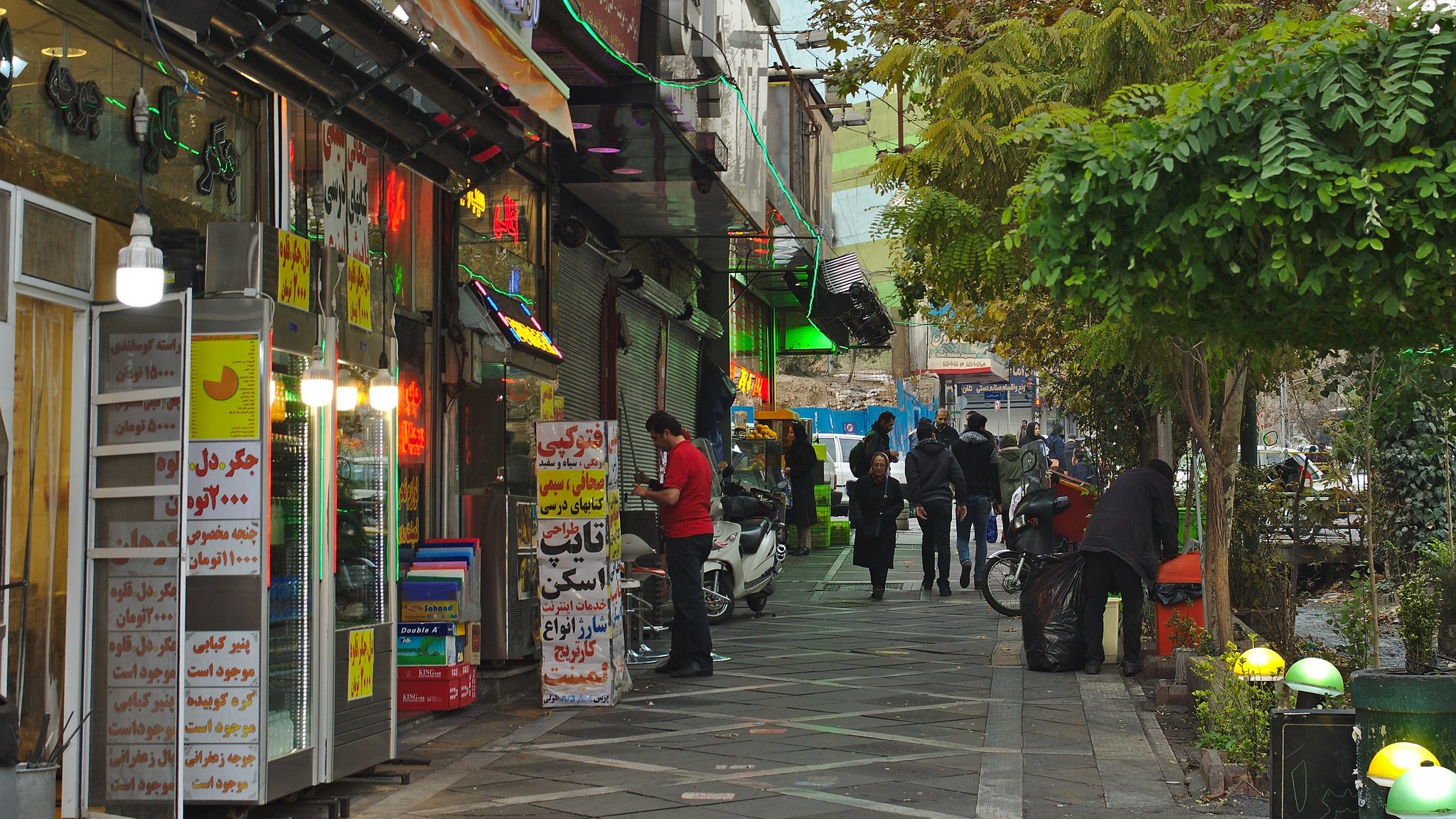 Une rue animée bordée de magasins colorés et de personnes marchant sous des arbres.
