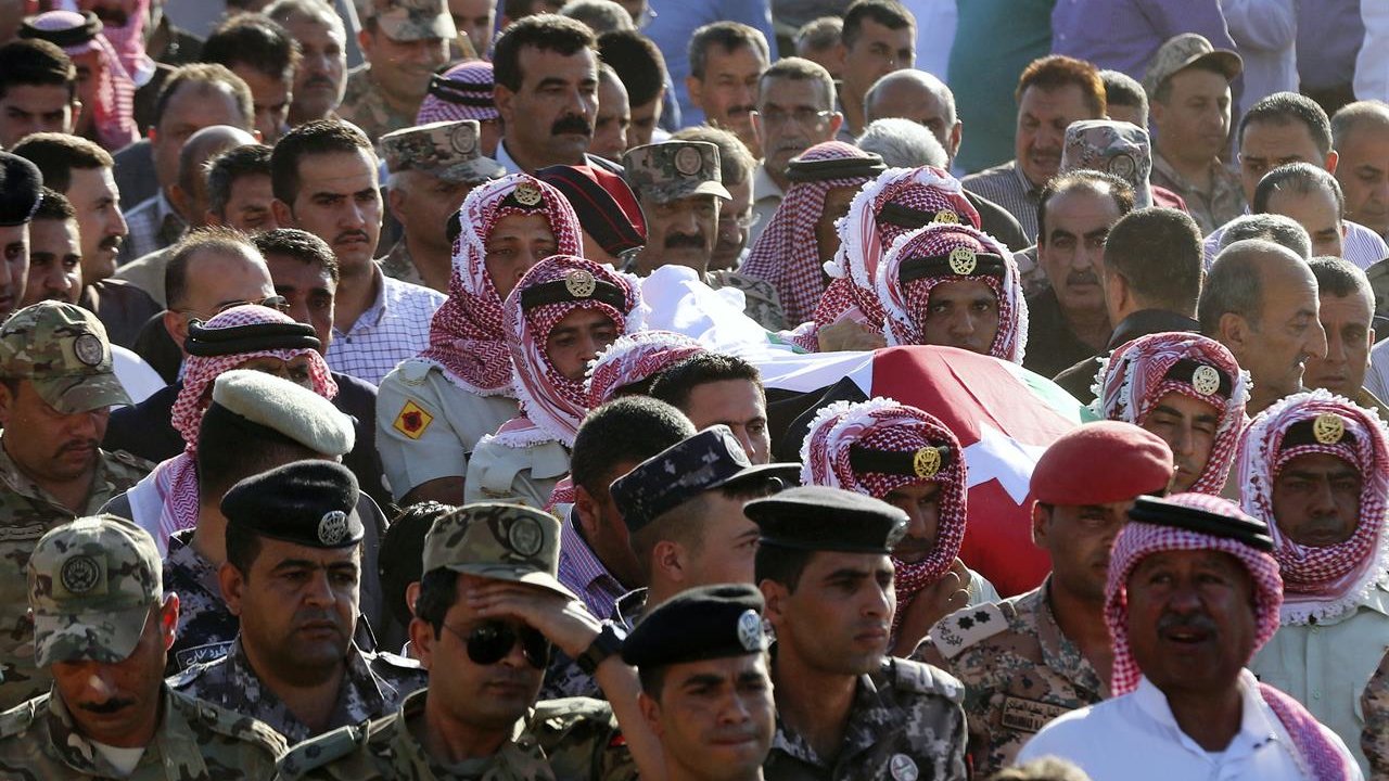 L'image montre une grande foule rassemblée. Au premier plan, des hommes portant des keffiehs (un type de couvre-chef traditionnel) entourent un cercueil recouvert d'un drapeau. Les participants semblent montrer leurs respects, et on peut également voir des militaires en uniforme dans la foule. L'atmosphère semble solennelle, indiquer une cérémonie ou des funérailles d'un individu important.