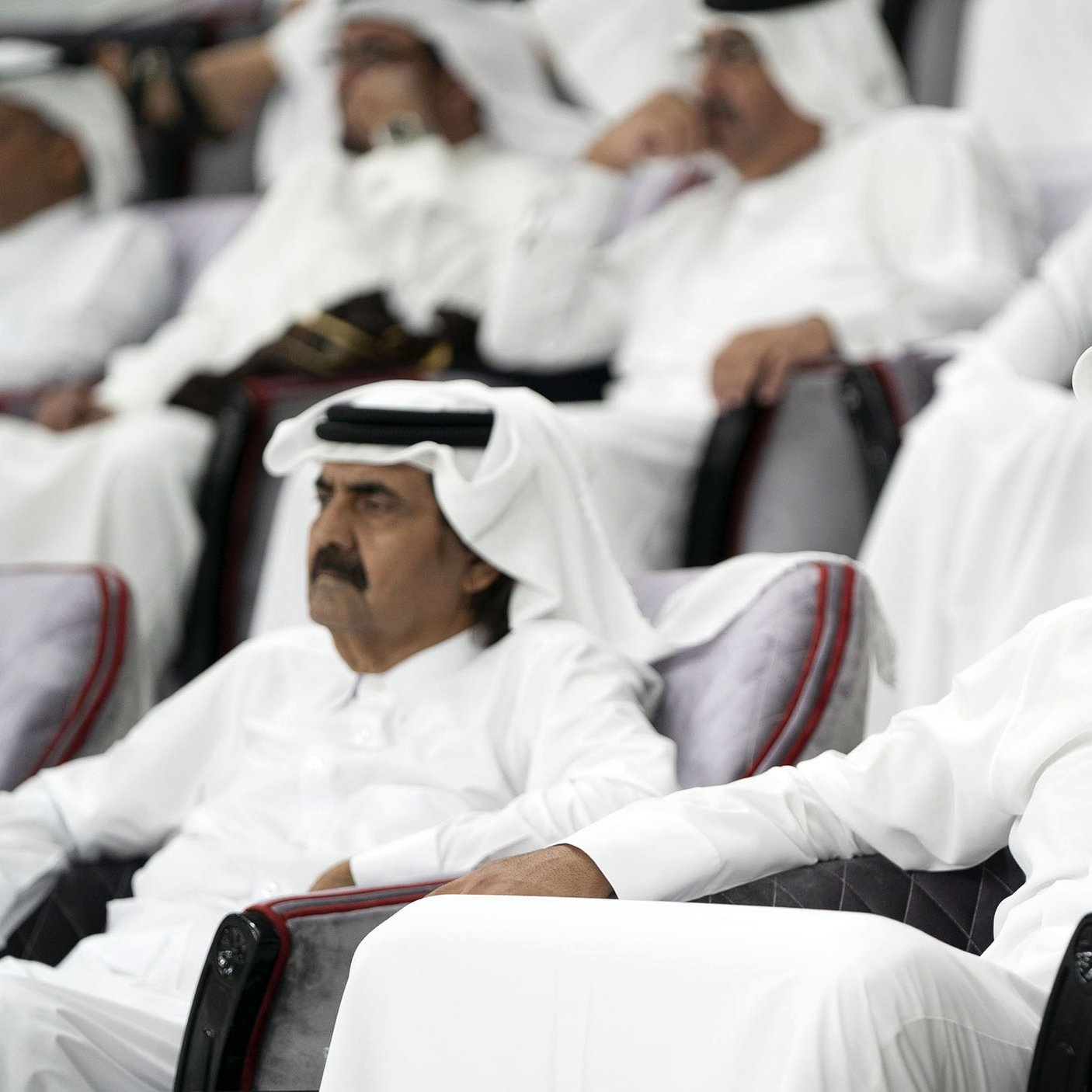 در این تصویر، دو مرد در حالی که لباس‌های سنتی عربی، شامل قمیص و عقال، پوشیده‌اند، نشسته‌اند. آن‌ها در یک مکان رسمی به تماشای چیزی مشغول‌اند. پس‌زمینه افراد دیگری را نشان می‌دهد که همچنین در لباس‌های سنتی نشسته‌اند. فضای کلی تصویر جدی و متمرکز به نظر می‌رسد.