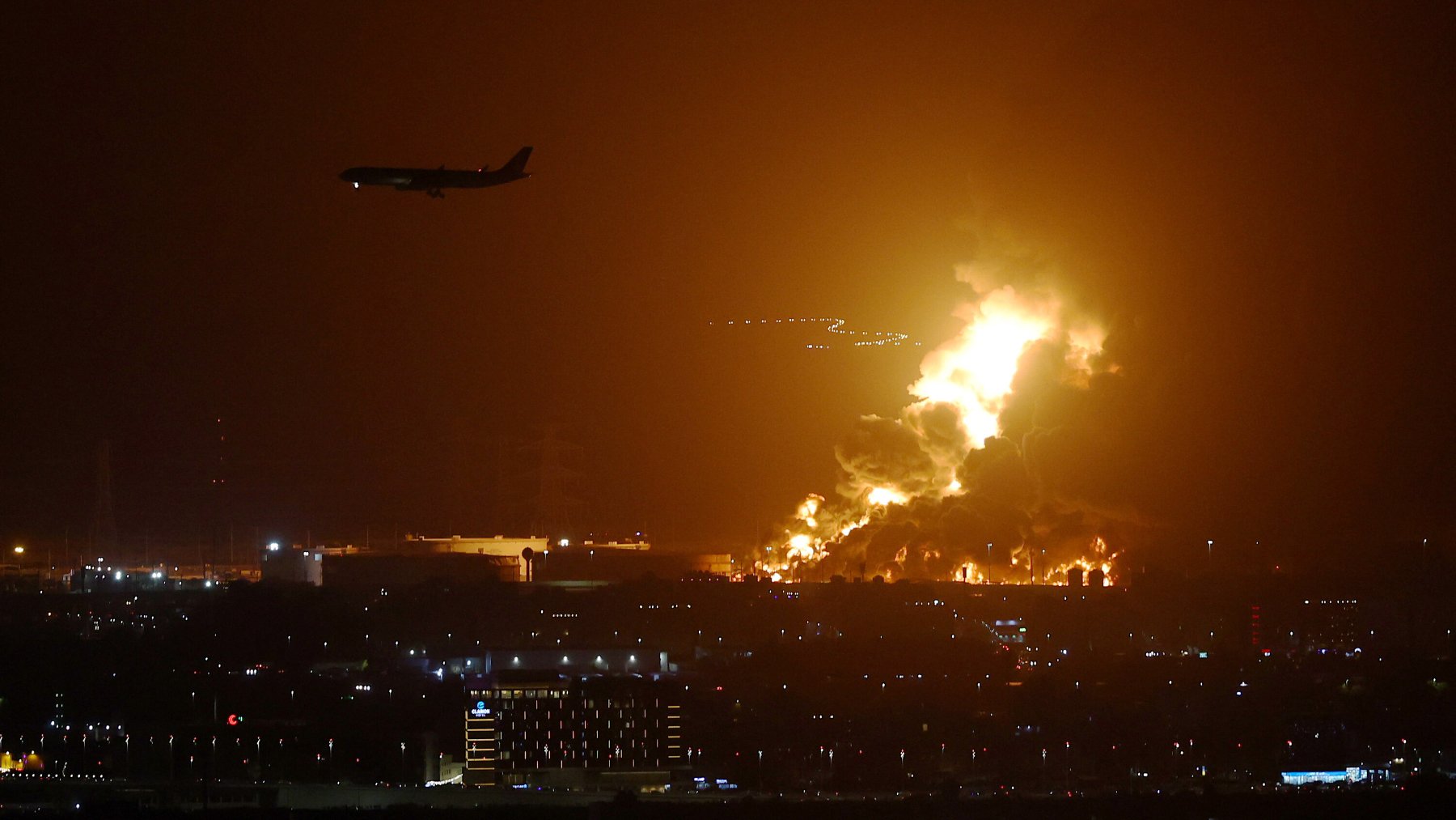 The image depicts a dramatic nighttime scene featuring a large, fiery blaze, likely arising from an industrial area. Bright flames and thick smoke illuminate the dark sky, creating a stark contrast against the surrounding darkness. In the foreground, there are city lights visible, indicating urban development, while an airplane can be seen flying overhead, possibly approaching a nearby airport. The scene suggests a serious incident, such as an explosion or fire, with significant visual impact.