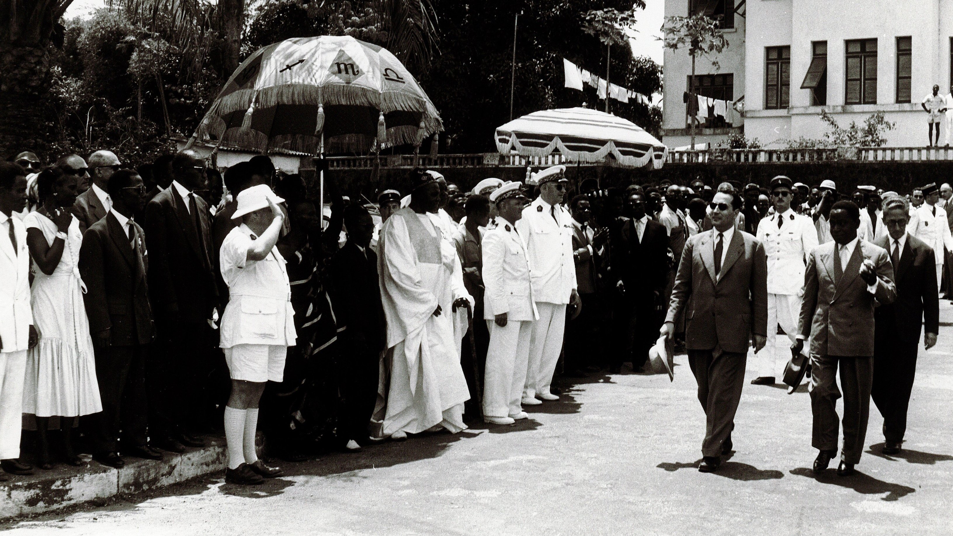 L'image montre une scène publique, probablement une cérémonie officielle. On y voit un groupe de personnes en costume, regroupées sur un côté, avec des hommes portant des chapeaux et des vêtements formels. À l'avant, on distingue des hommes en uniformes militaires et d'autres en tenue élégante. Au centre, une figure principale semble marcher vers la caméra, entourée de gardes et d'accompagnateurs. Des parasols colorés sont également visibles, ajoutant une touche d'ombre à la scène. En arrière-plan, on peut apercevoir un bâtiment officiel avec des drapeaux, indiquant un événement important ou une célébration. L'atmosphère semble solennelle et organisée.