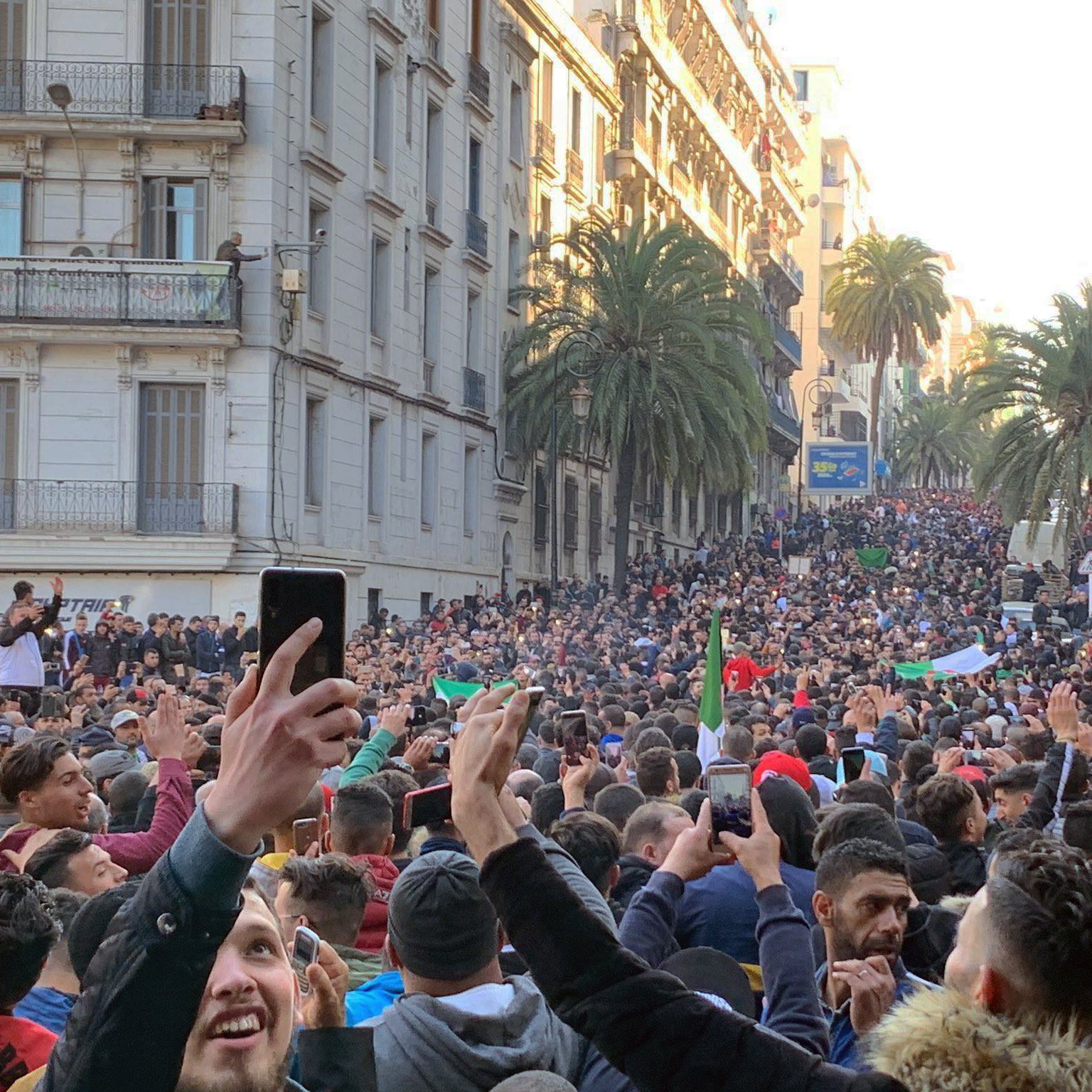 این تصویر نمایی از یک اجتماعات بزرگ در خیابانی شلوغ را نشان می‌دهد. مردم زیادی در حال تظاهرات یا جمع شدن هستند و در میان آن‌ها تعدادی پرچم به چشم می‌خورد. درختان بلند نخل در دو طرف خیابان دیده می‌شوند و ساختمان‌های مرتفع با بالکن‌ها نیز در پس‌زمینه قرار دارند. مردم با entusiasmo در حال برنداشتن تلفن‌های همراه خود و ثبت این لحظه هستند. جو عمومی تصویری از شور و شوق و همبستگی را القا می‌کند.