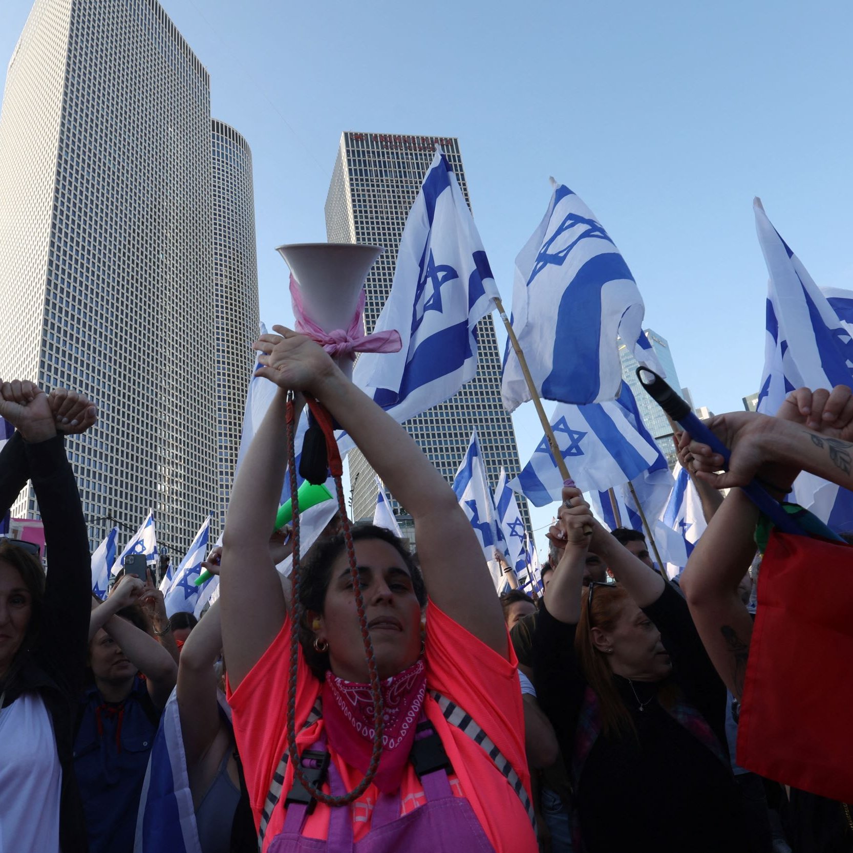 تظهر الصورة مجموعة من الأشخاص في مظاهرة، يحملون أعلامًا تحمل نجمة داود، وهو رمز معروف في إسرائيل. تظهر امرأة في المركز، مرفوعة اليدين وتحمل جهاز مكبر صوت، بينما يرفع الآخرون الأعلام. تتواجد خلفهم بنايات حديثة، مما يشير إلى أن المظاهرة قد تكون في منطقة حضرية. يعبّر المتظاهرون عن آرائهم، مما يدل على أهمية الفعالية التي يشاركون فيها.